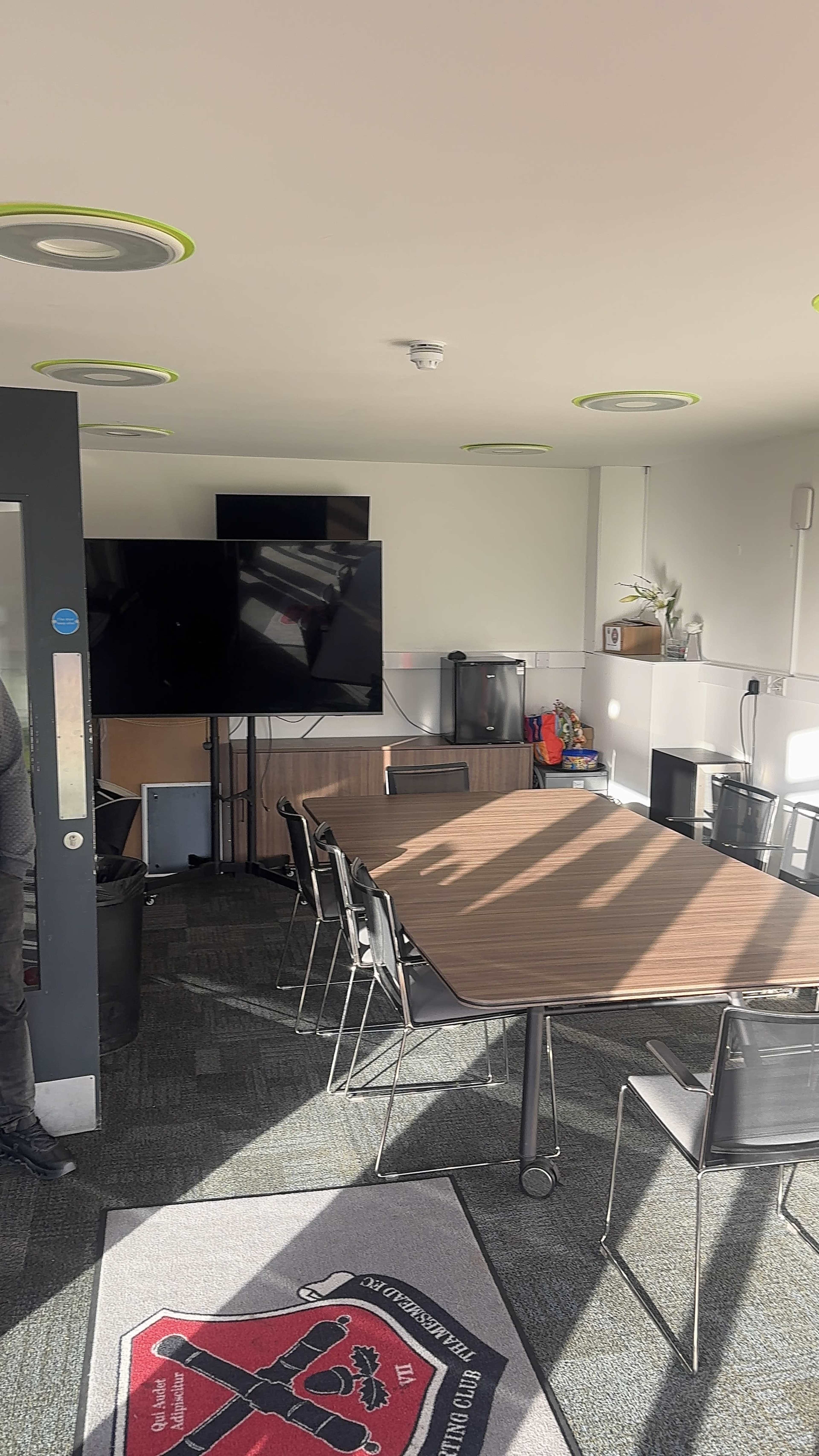 The image shows a meeting room with a large table, chairs, and two television screens mounted on the wall.