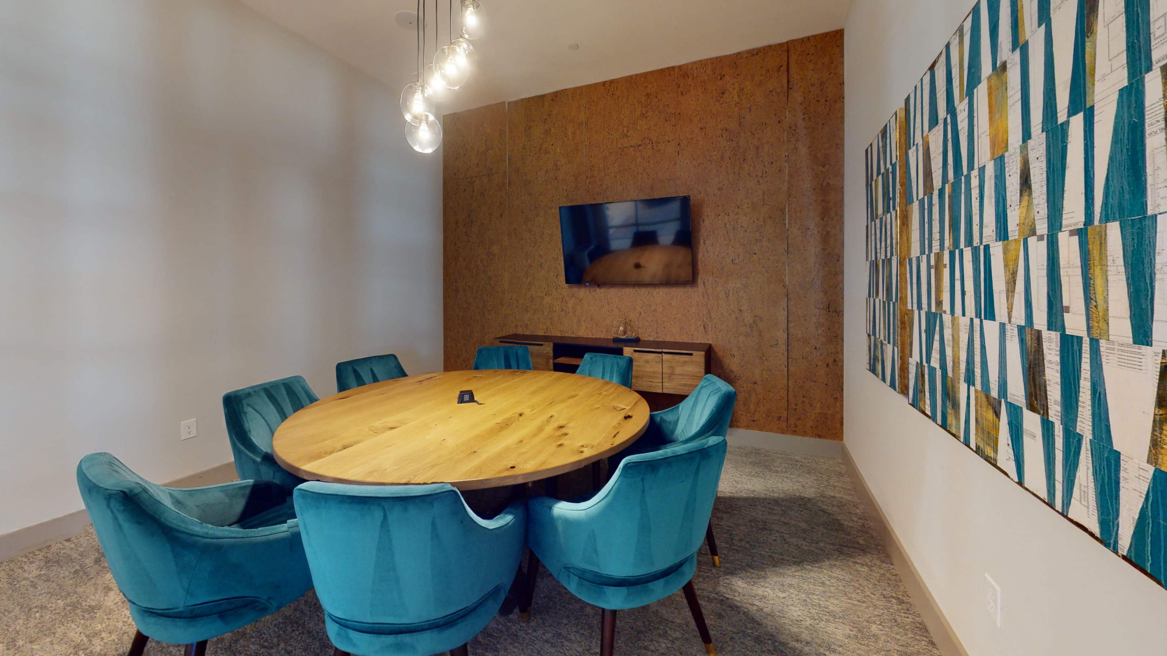 A modern conference room featuring a round wooden table surrounded by teal upholstered chairs, with a wall-mounted TV and artwork in the background.