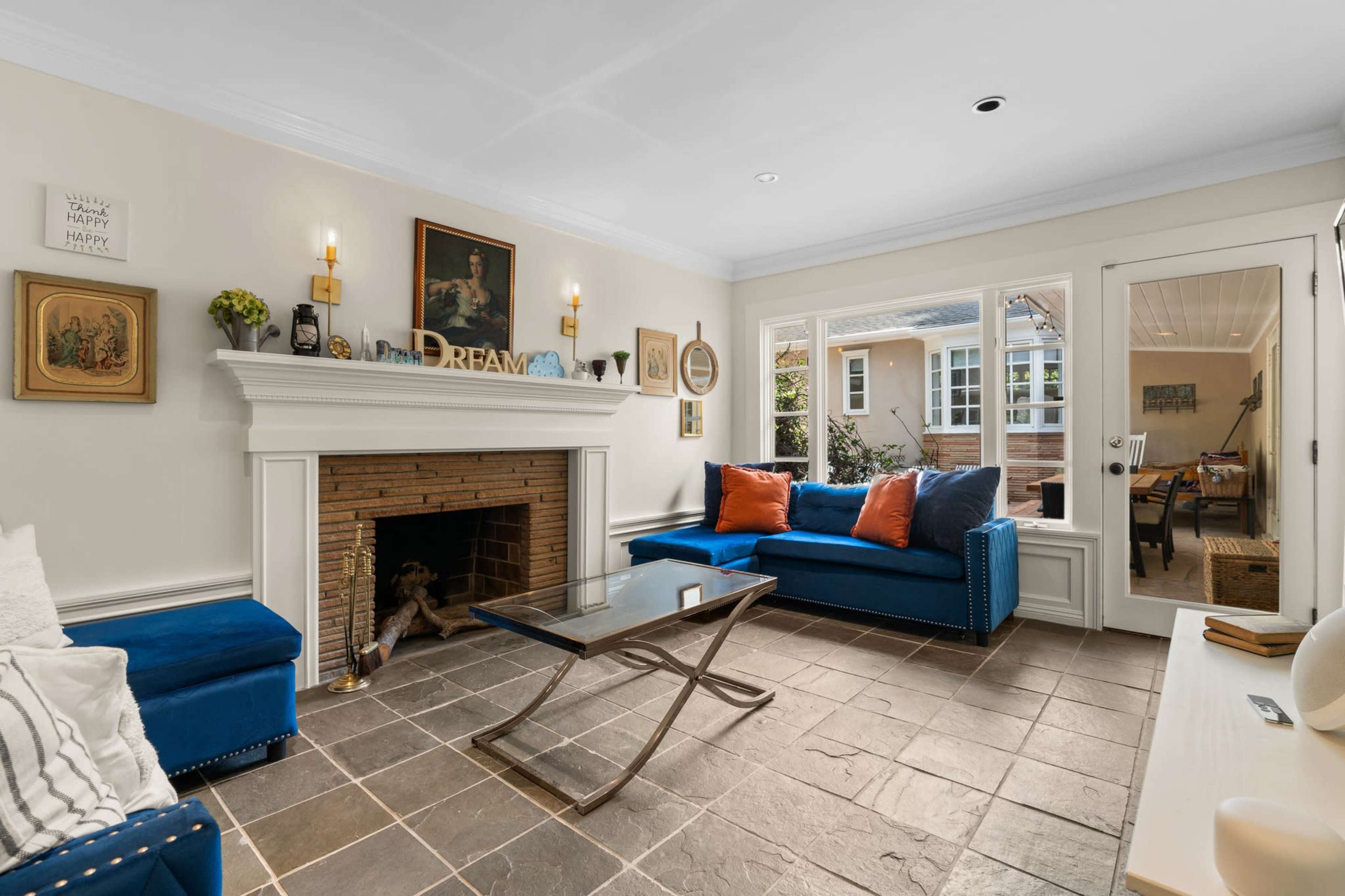 The living room features a brick fireplace, a blue sofa with orange pillows, and a glass coffee table, with large windows allowing light to enter from an adjoining room.