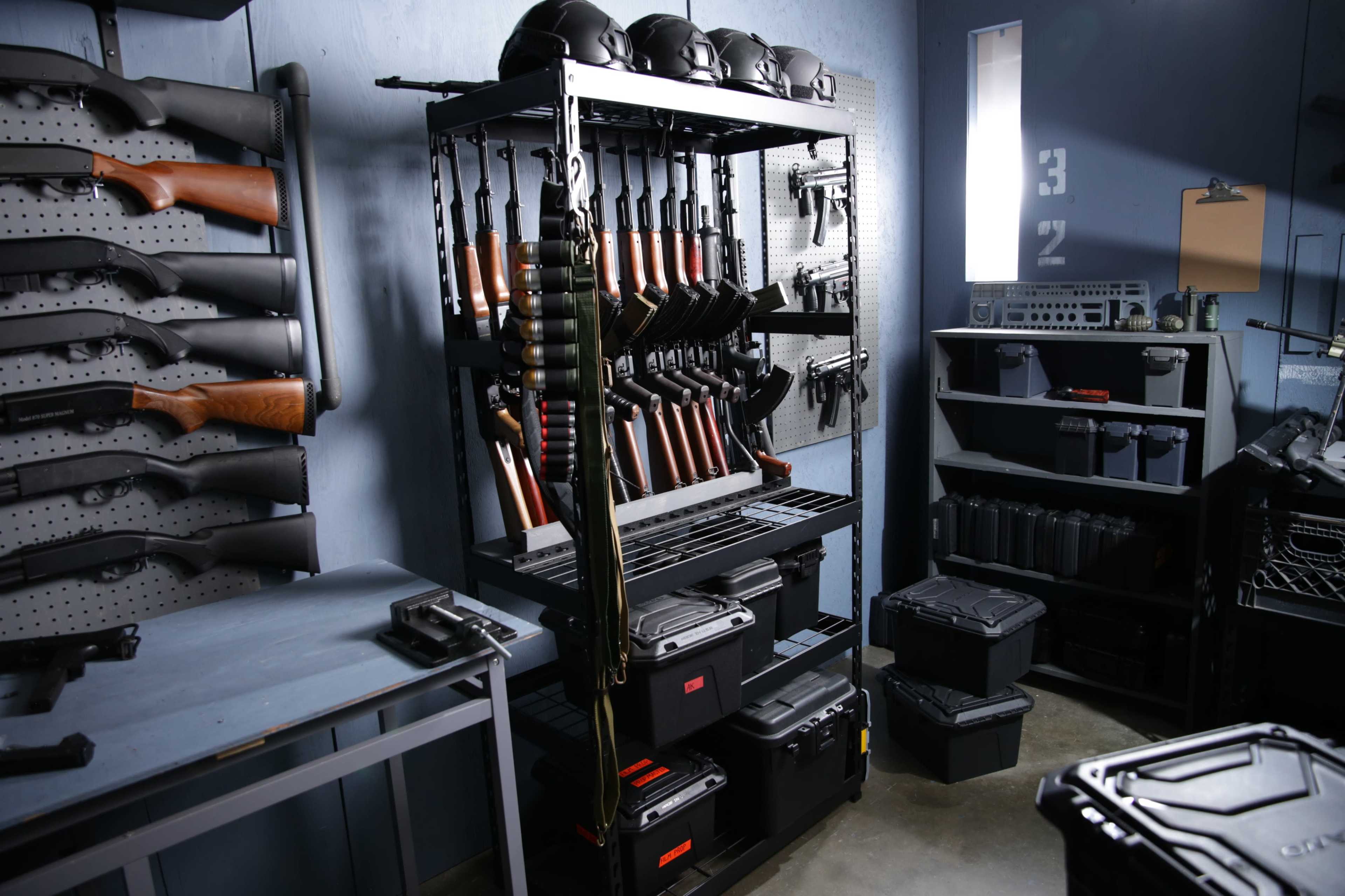 The image shows a storage area equipped with various firearms mounted on racks, tactical helmets on top, and ammunition crates on the floor.