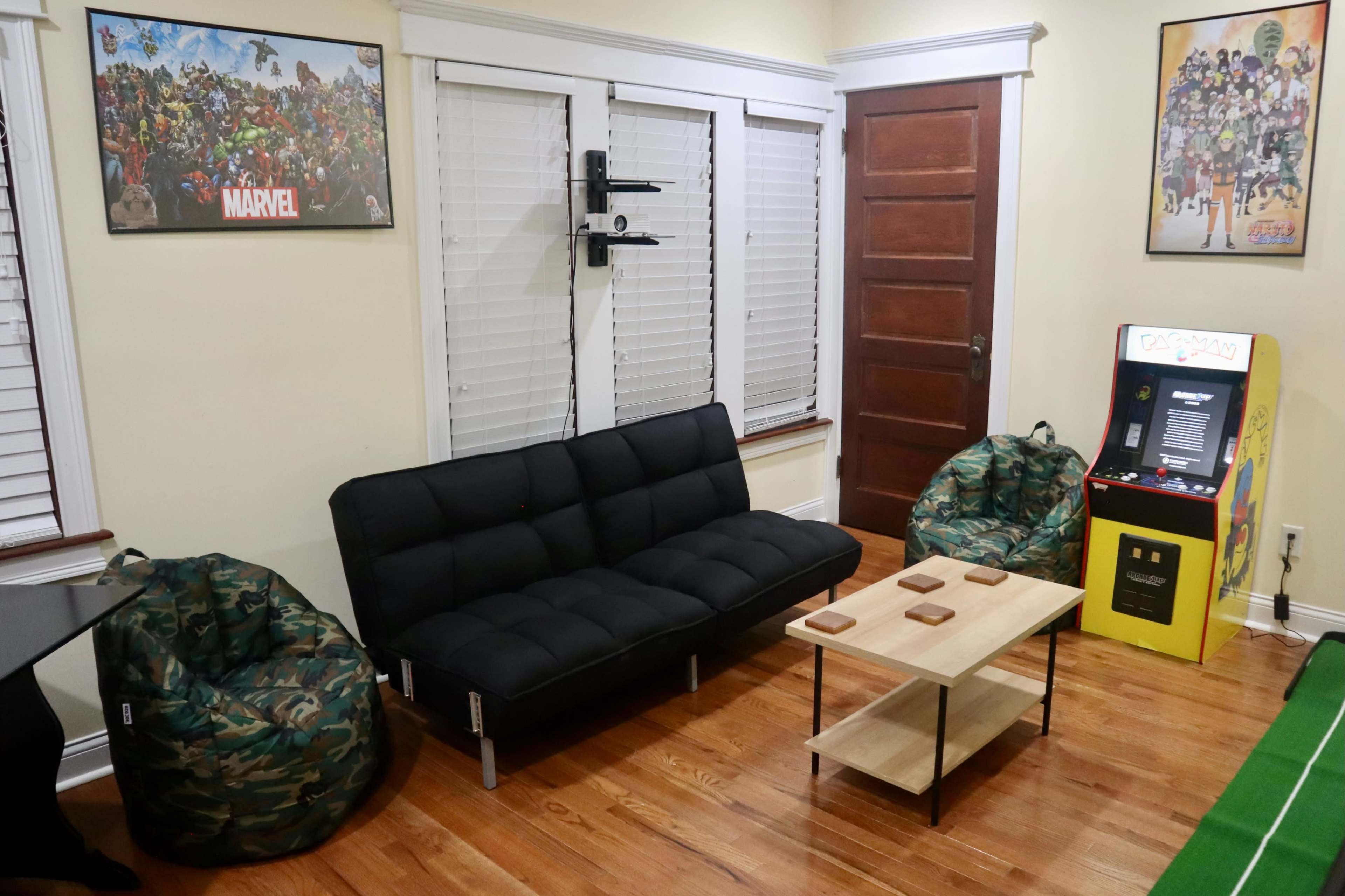 A living room featuring a black sofa, two camouflaged bean bags, a wooden coffee table, and a vintage arcade game machine in a brightly colored cabinet.