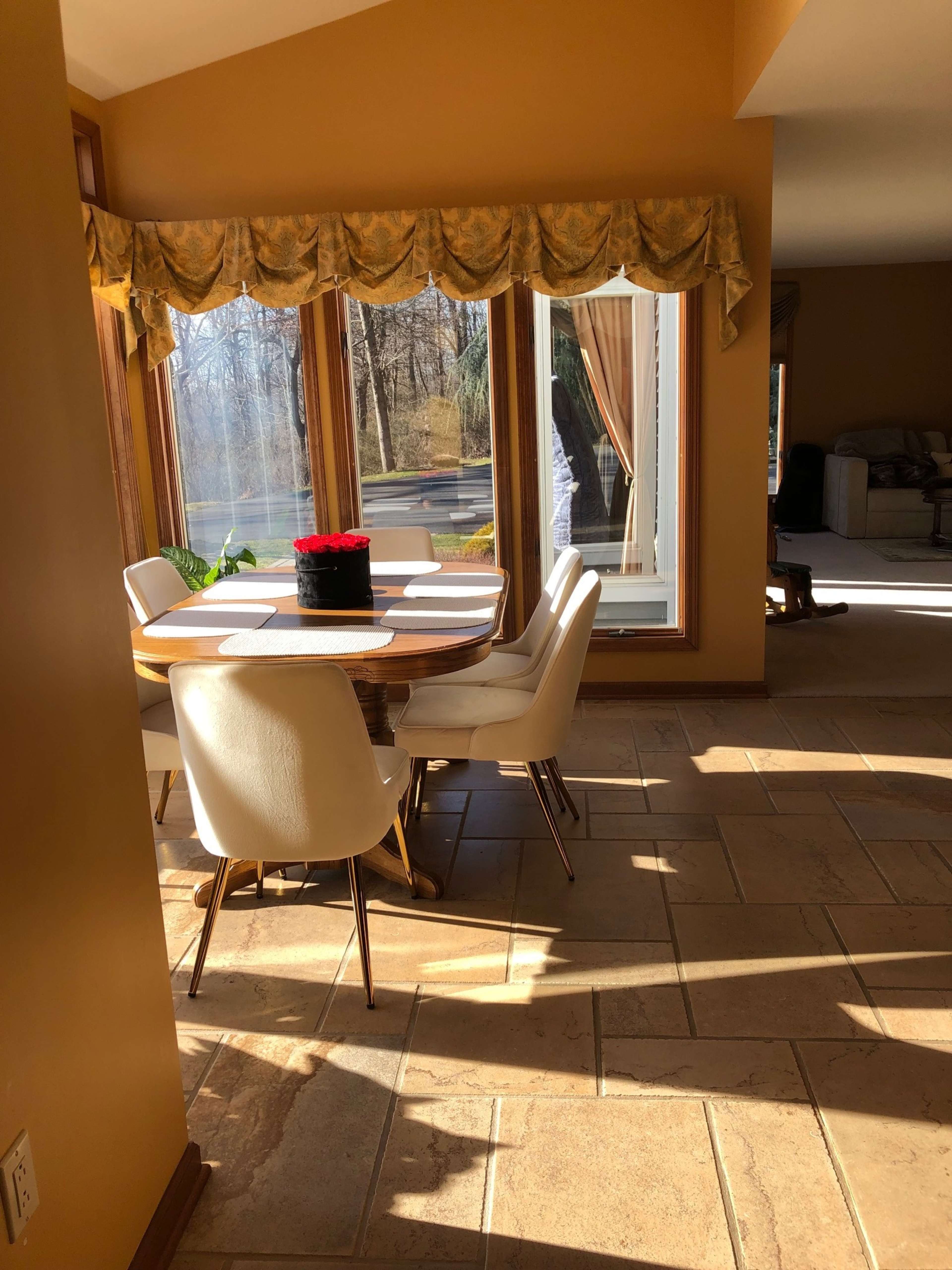 A dining area with a round wooden table and white chairs is illuminated by sunlight streaming through large windows.