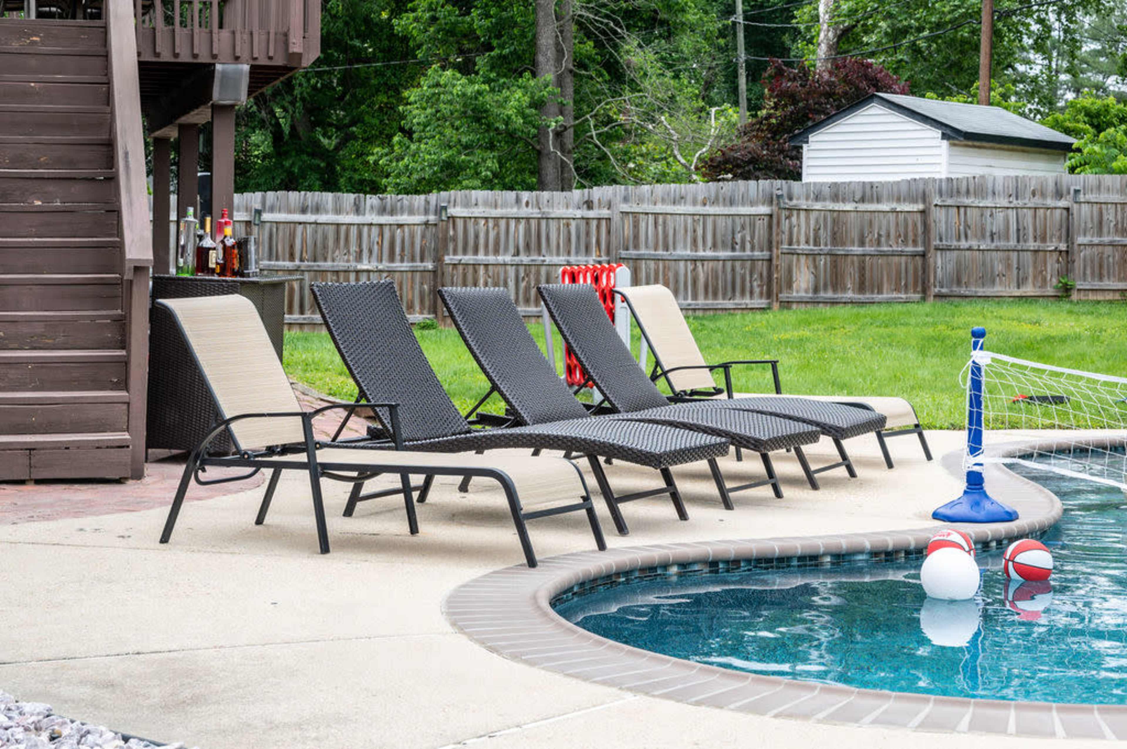 A set of four lounge chairs lined up by a swimming pool, with a couple of beach balls floating nearby.