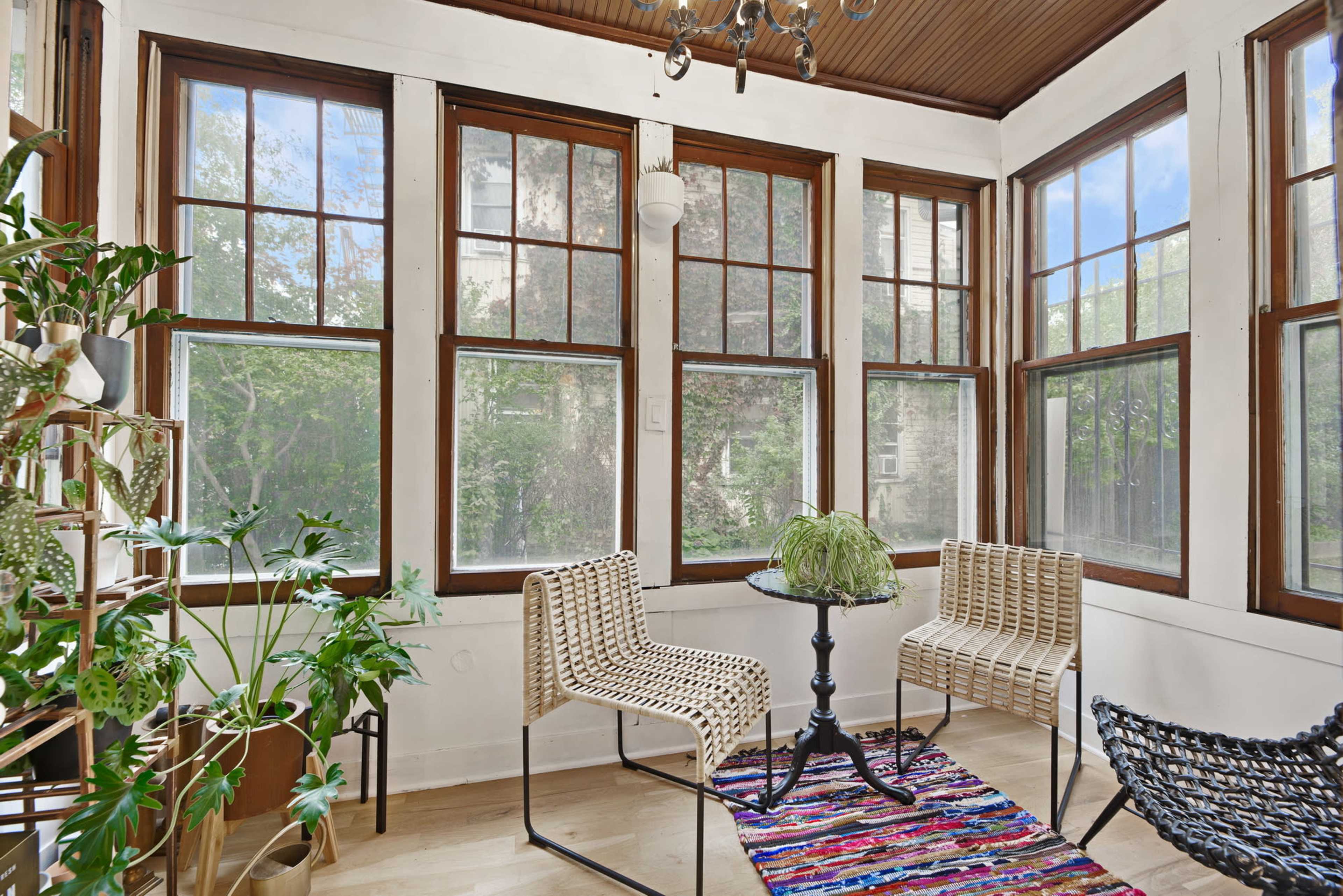 A sunlit sunroom features large windows, a small round table, and wicker chairs surrounded by indoor plants.