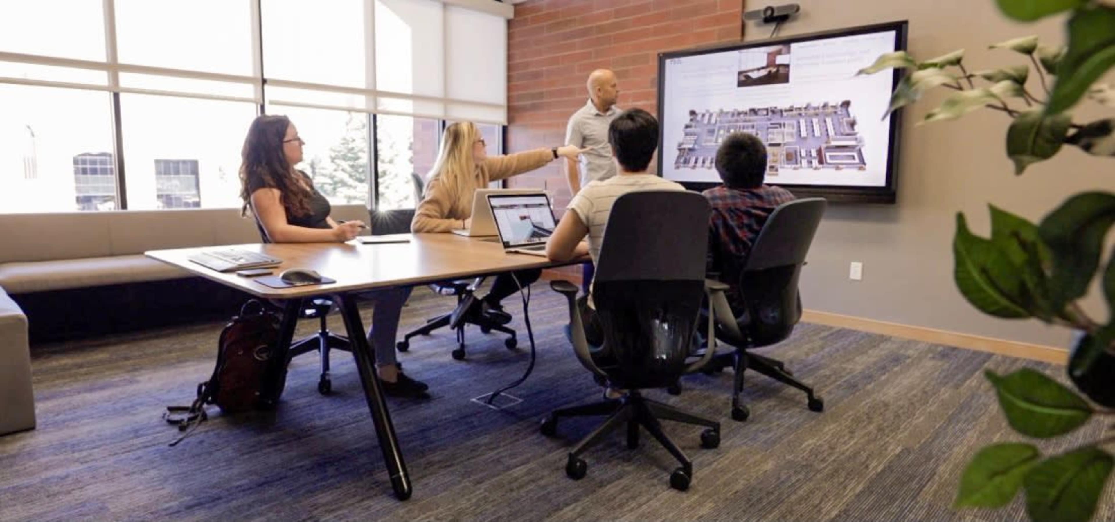 A group of four people are seated around a table in a conference room, while one person stands and presents information on a large screen.
