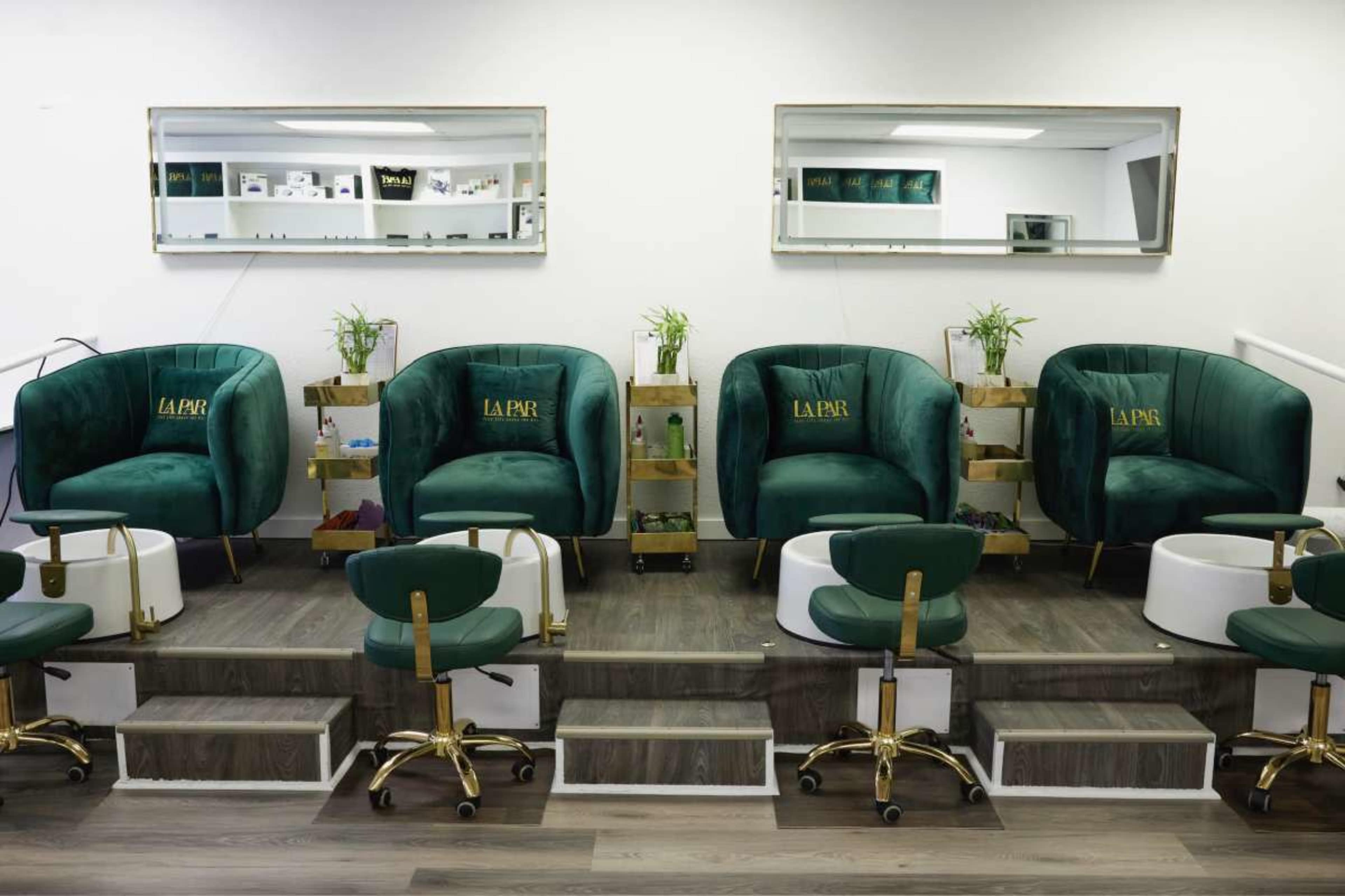 The image shows a nail salon with four plush green chairs arranged in a row, each paired with a foot bath and decorated with plants, while mirrors reflect the space above them.