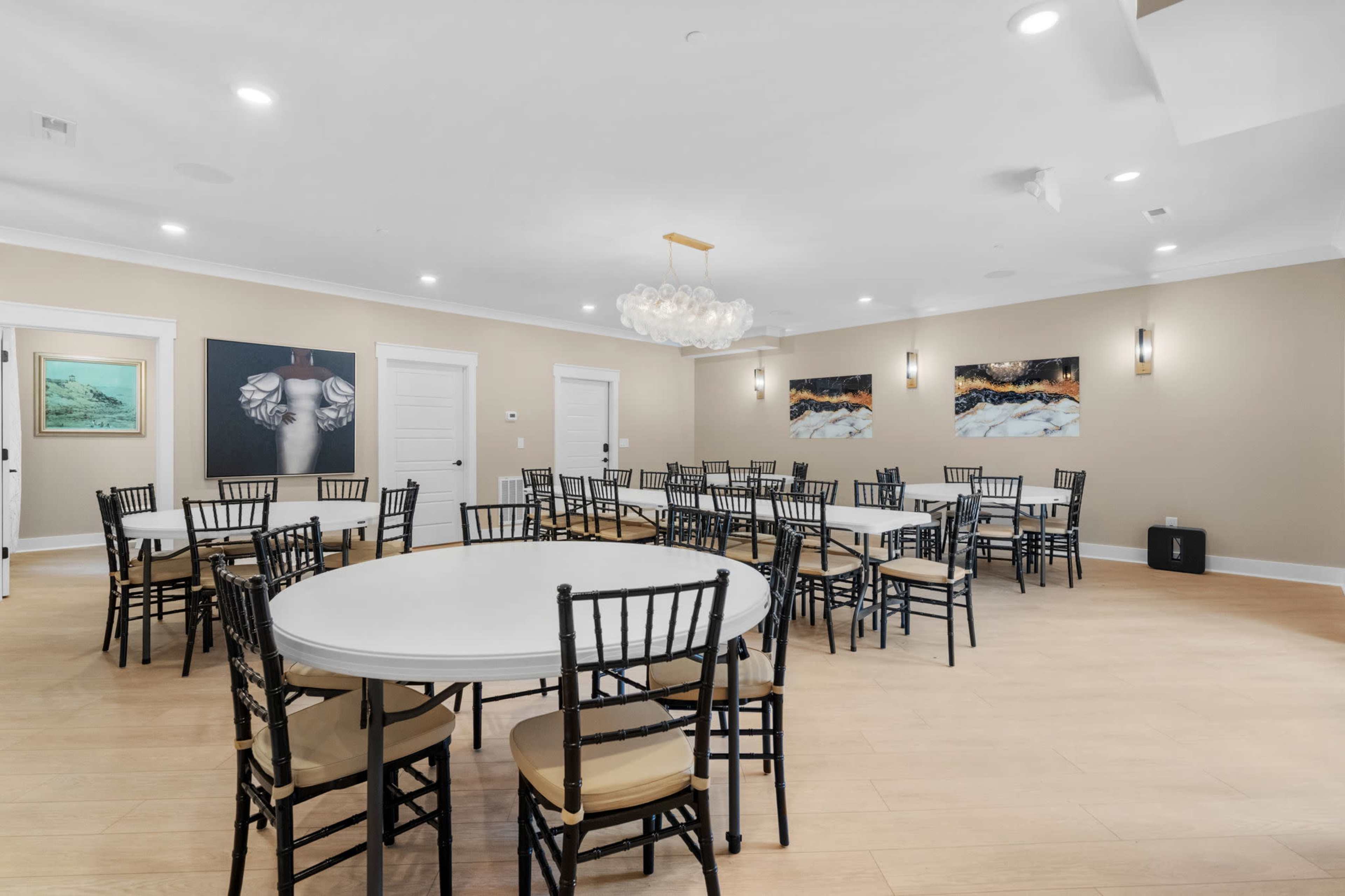 The image shows a well-lit event room with multiple round tables set with black chairs, featuring artwork on the walls and a chandelier overhead.
