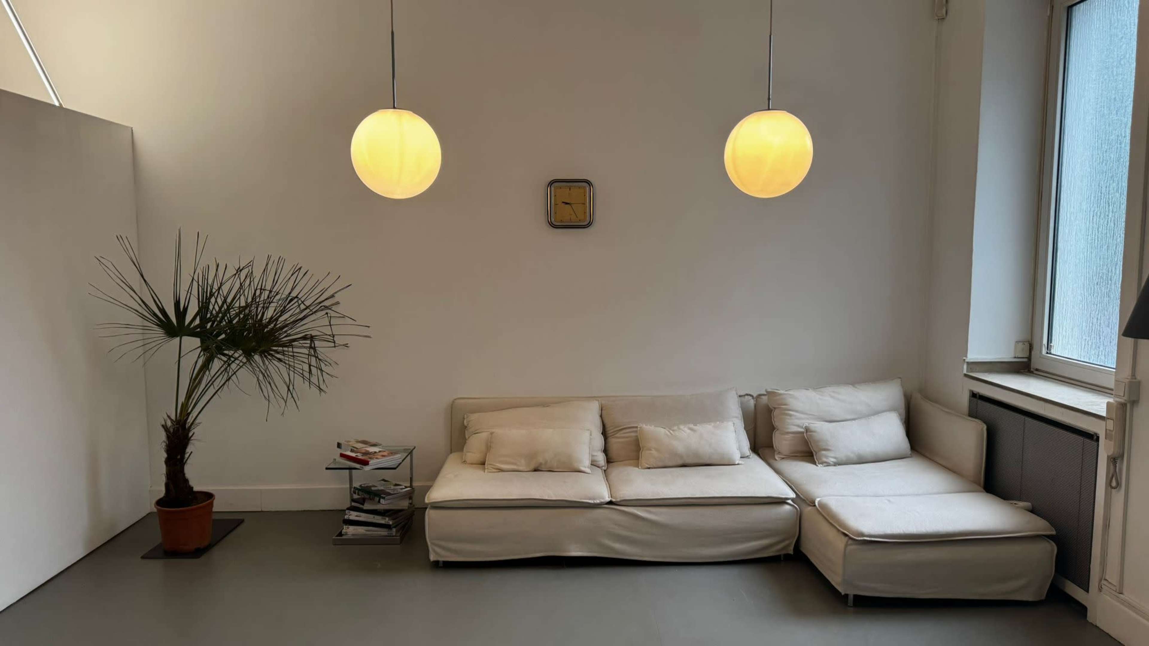 The image shows a minimalist living space featuring a light-colored sectional sofa, a potted palm plant, and two round pendant lights hanging from the ceiling.