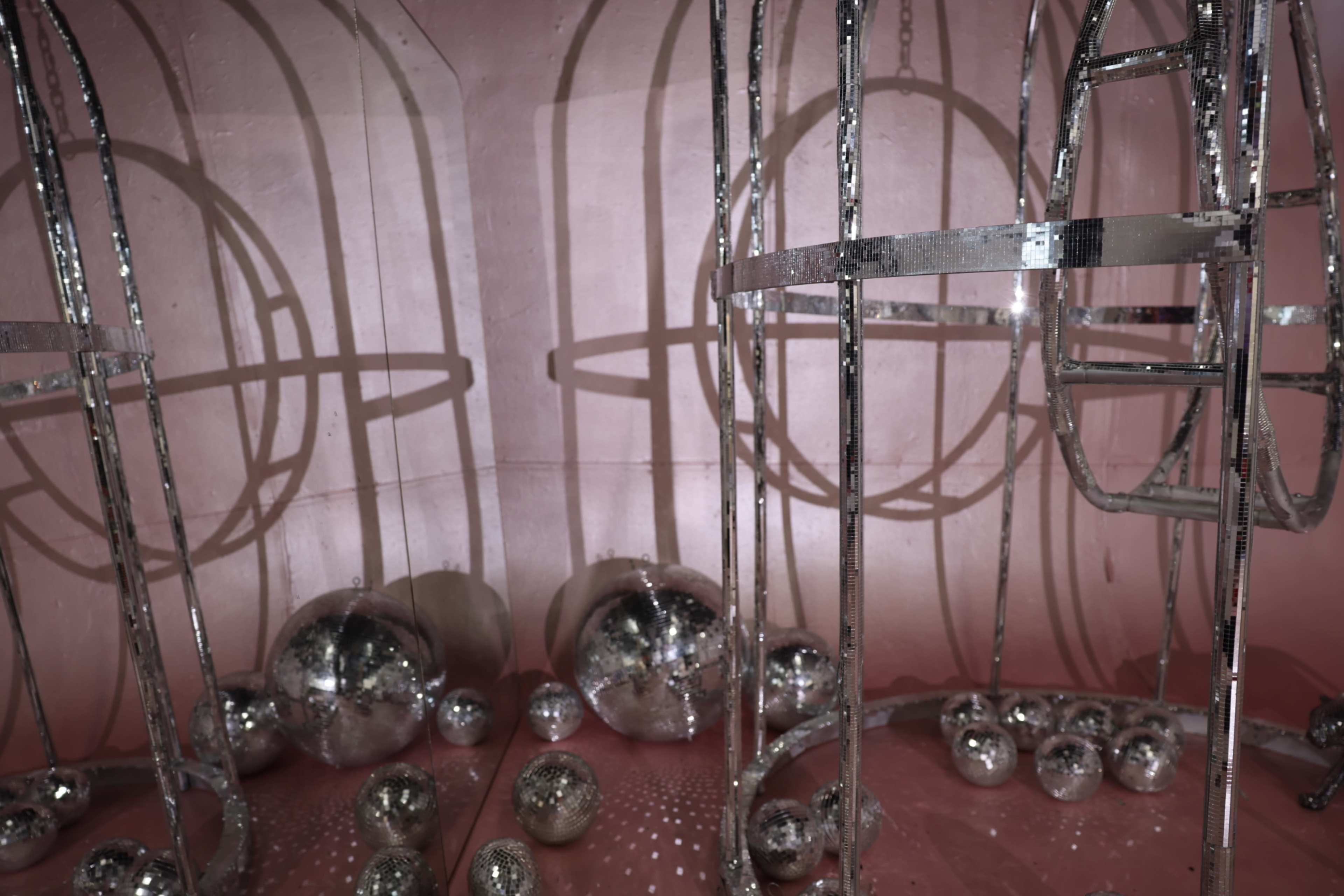 The image features a pink interior with metallic structures casting circular shadows and a collection of reflective spheres scattered on the floor.
