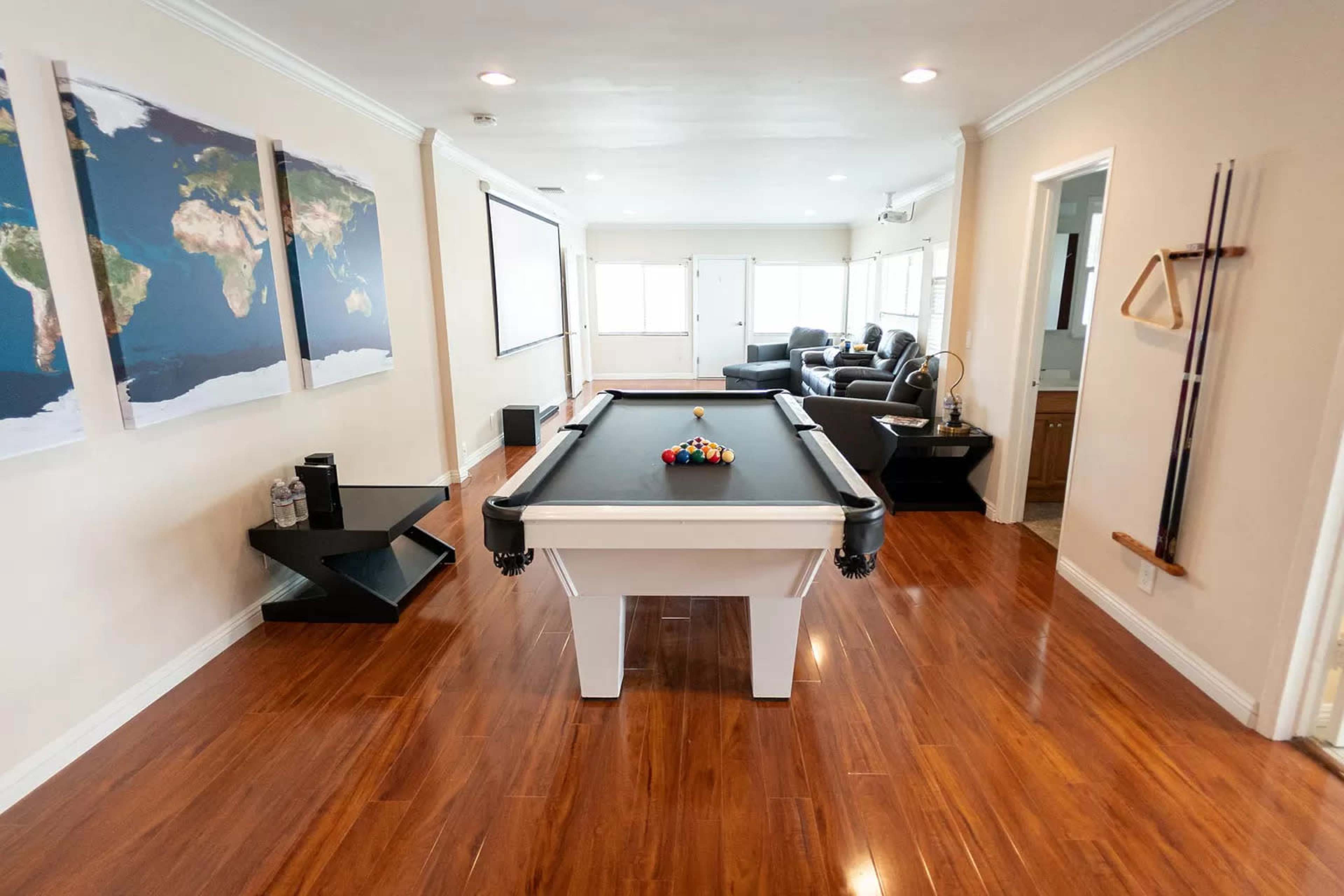 The image shows a spacious room featuring a pool table in the center, with a world map on the wall and a seating area visible in the background.