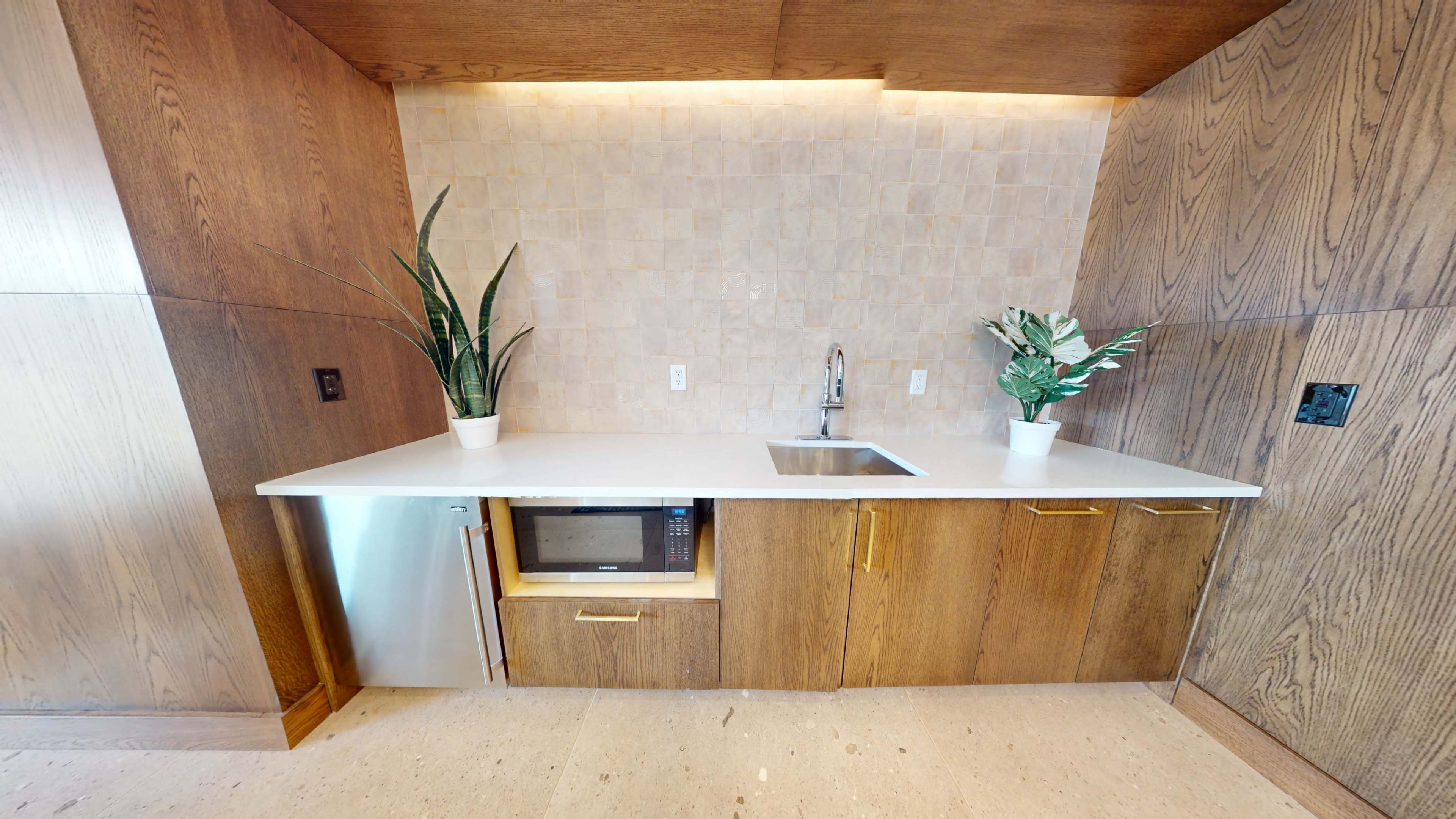 A modern kitchen area features a sink, microwave, and tall plants on a countertop with wooden cabinetry and a tiled backsplash.