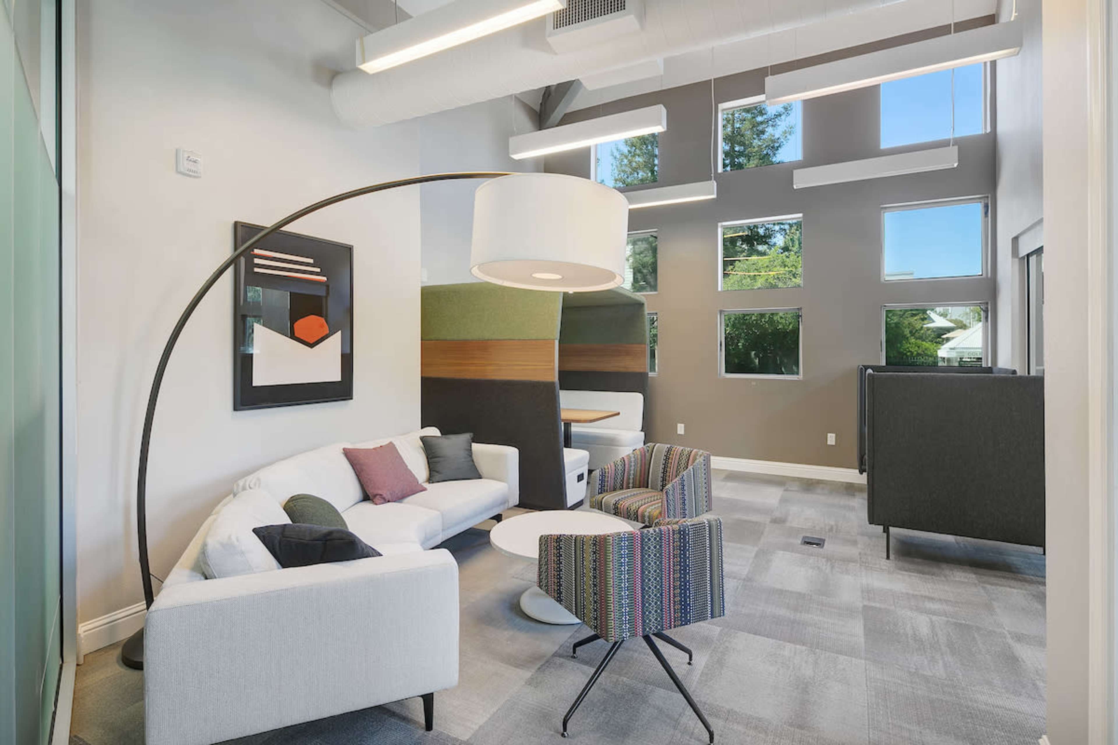 The image shows a modern seating area with a curved lamp, a white couch, a colorful patterned chair, a round table, and large windows providing natural light.