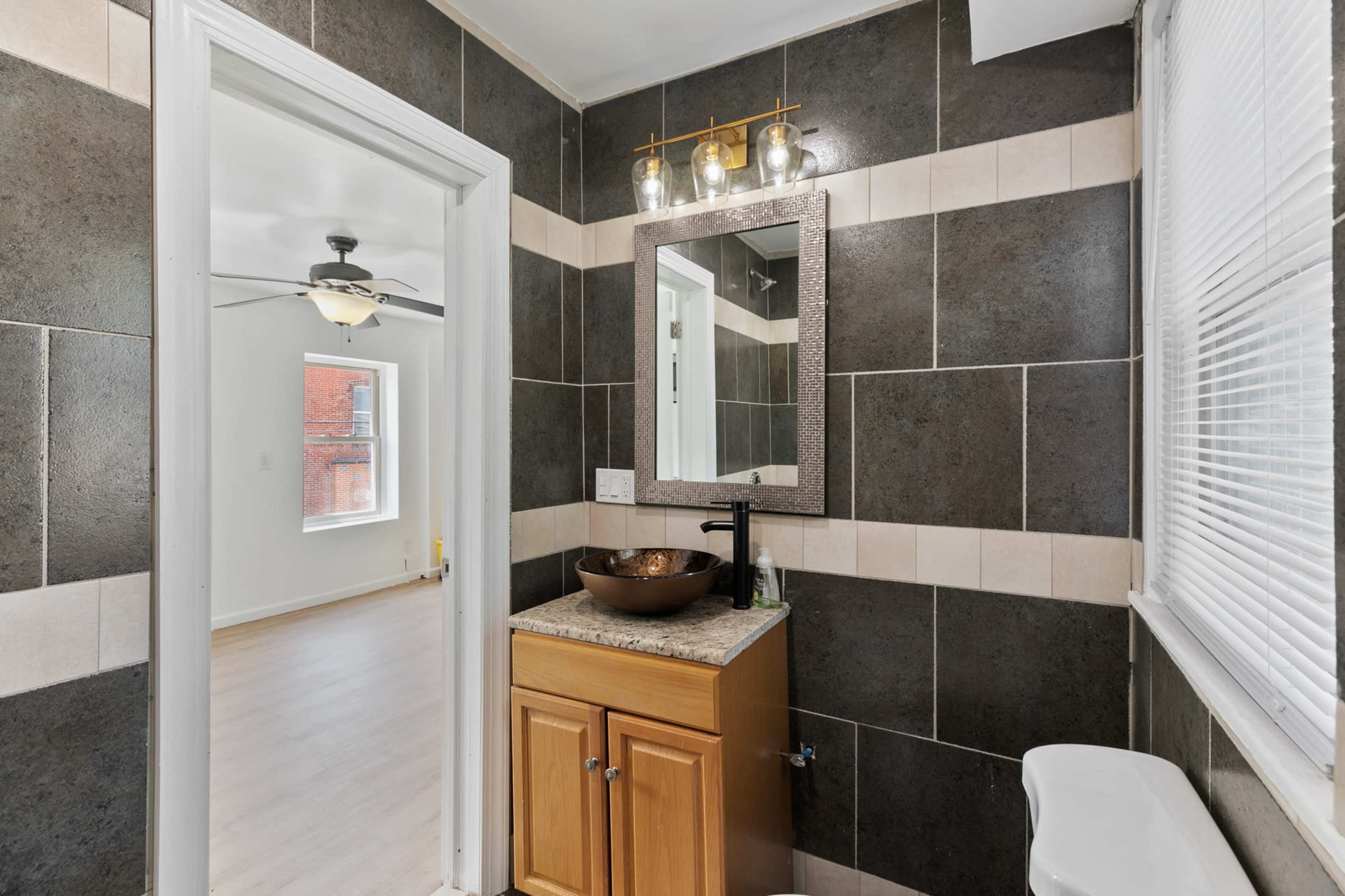 A modern bathroom features dark tiled walls, a wooden cabinet with a round bowl sink, and a mirror with a light fixture above it.