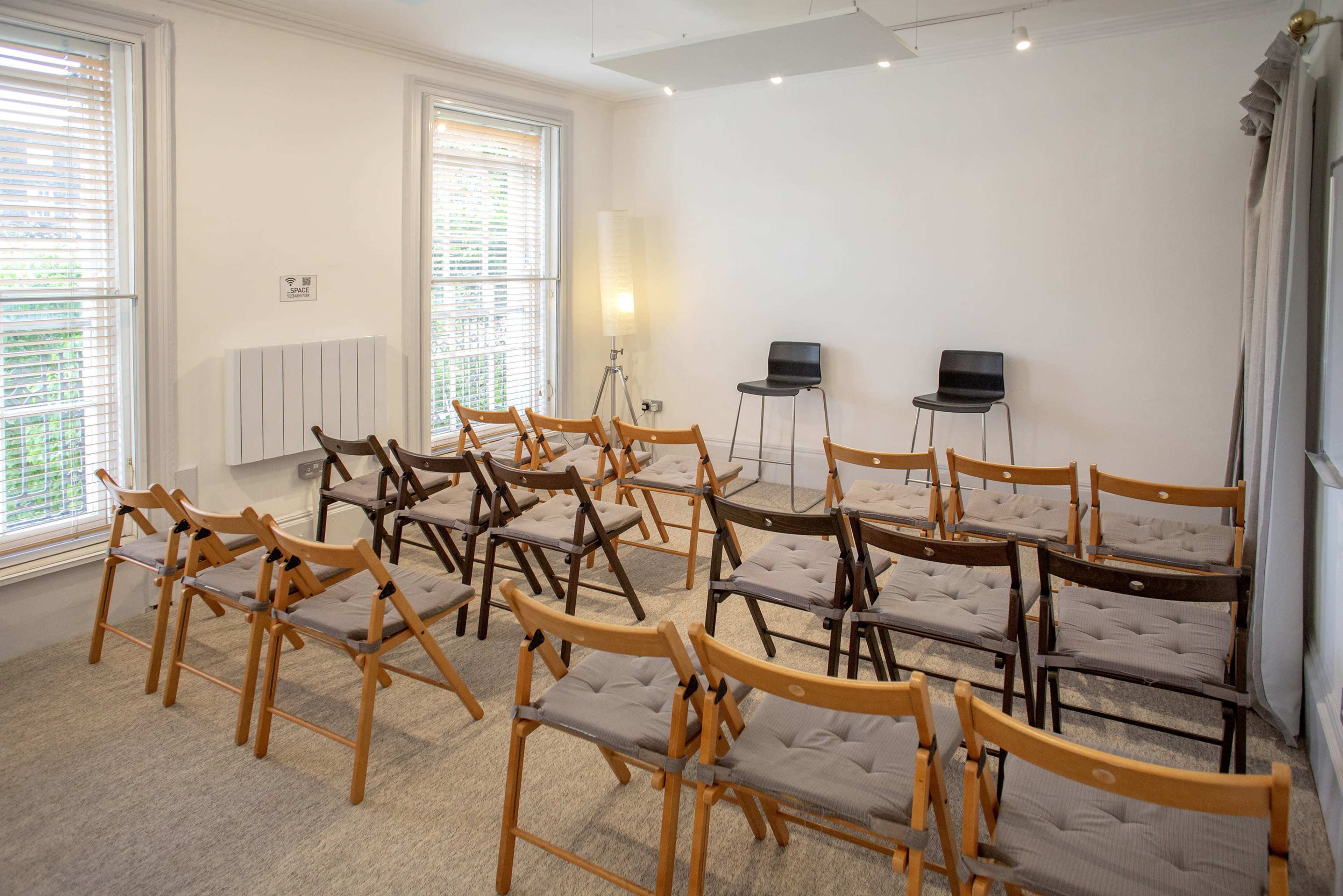 The image shows a room with rows of wooden folding chairs arranged facing two black chairs at the front.