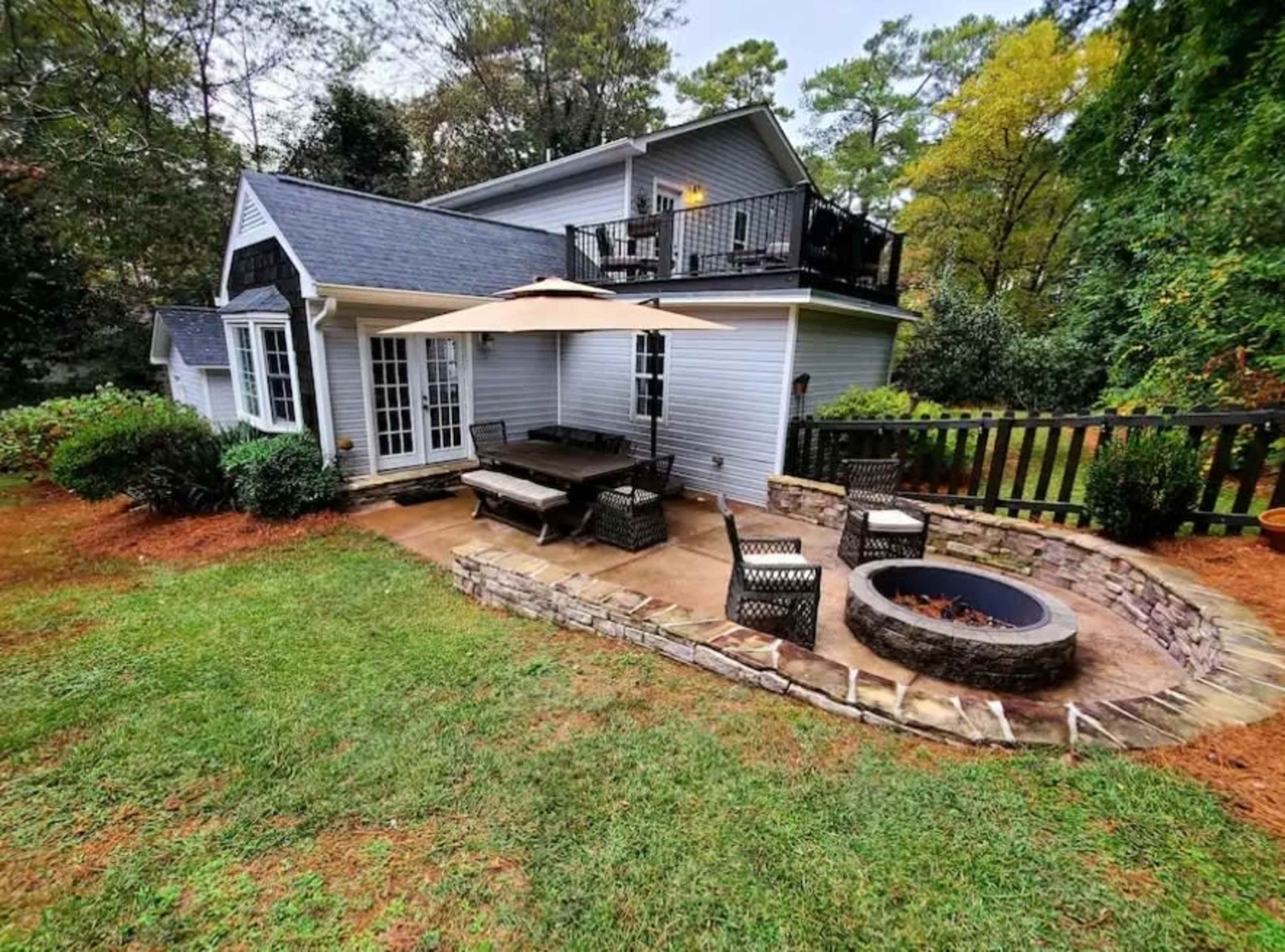 The image shows a backyard patio with a stone fire pit, seating area, and a shaded dining table next to a house surrounded by greenery.