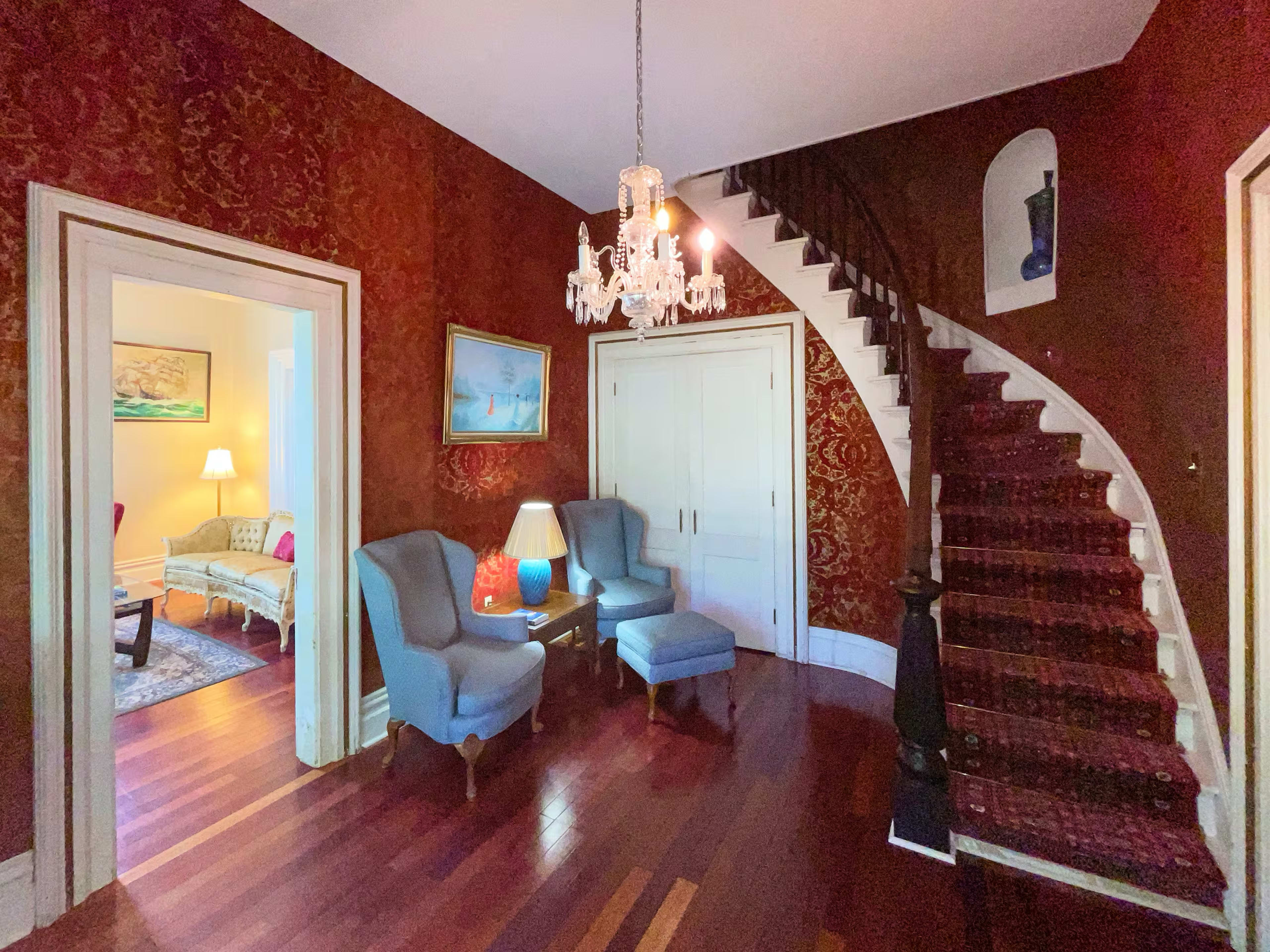 The image shows a hallway with a staircase, featuring ornate wallpaper, two blue chairs with a side table, and an adjacent room visible in the background.