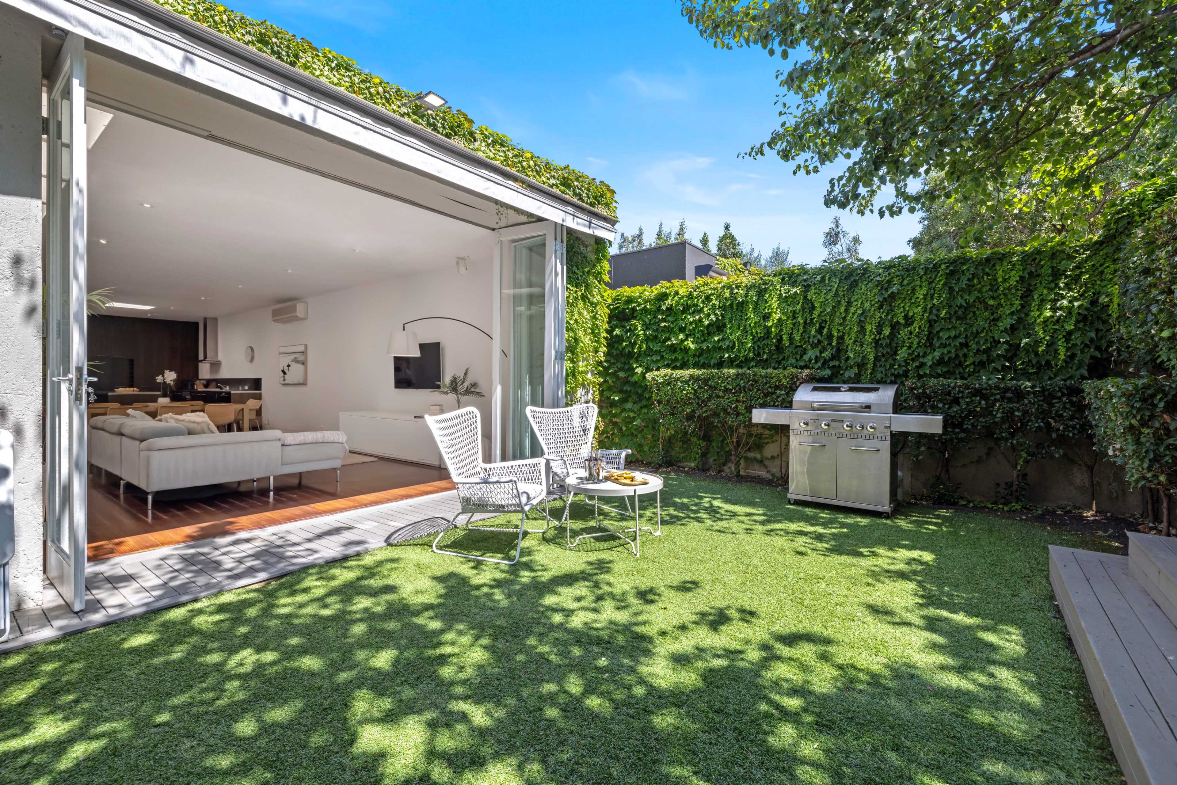A modern backyard features two chairs, a small table, and a stainless steel grill surrounded by lush green grass and hedges.
