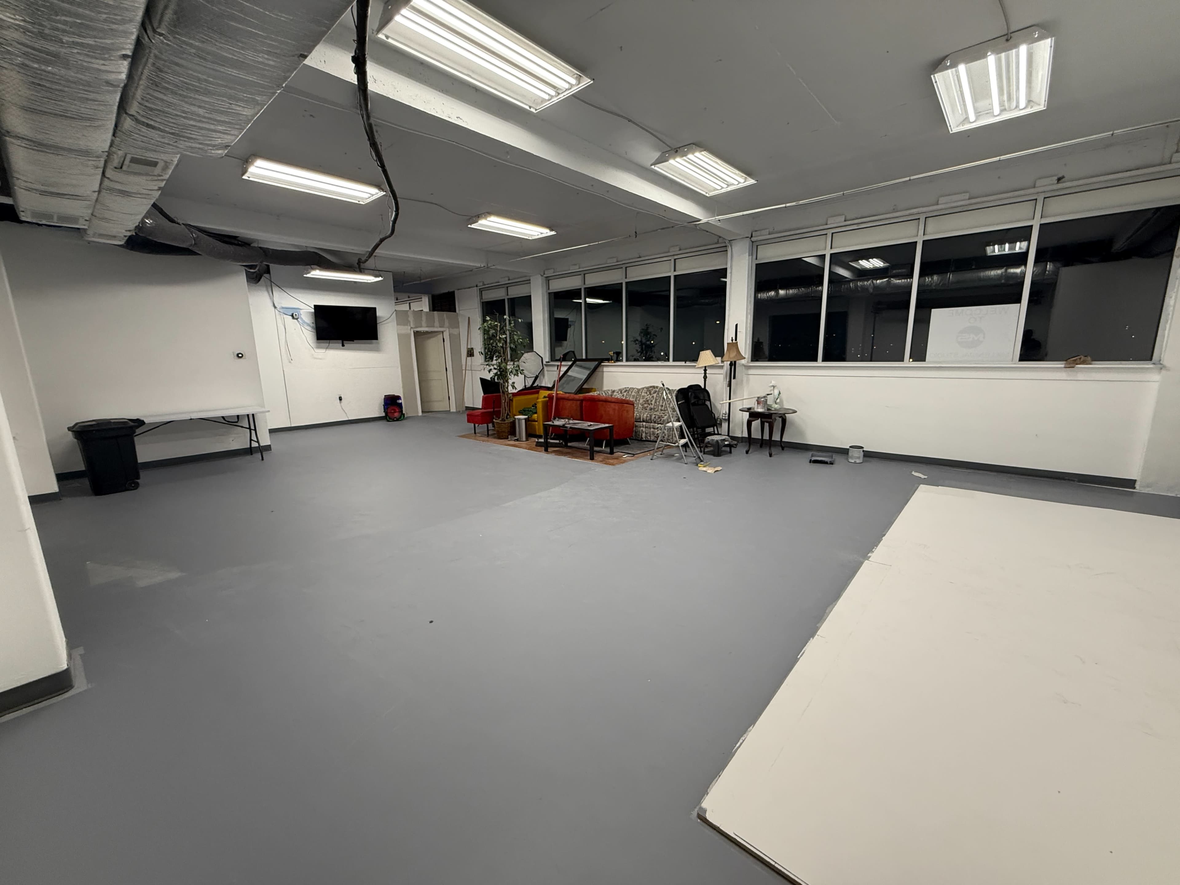 The image shows an empty, spacious room with gray flooring, large windows, and minimal furniture including a sofa and a table.