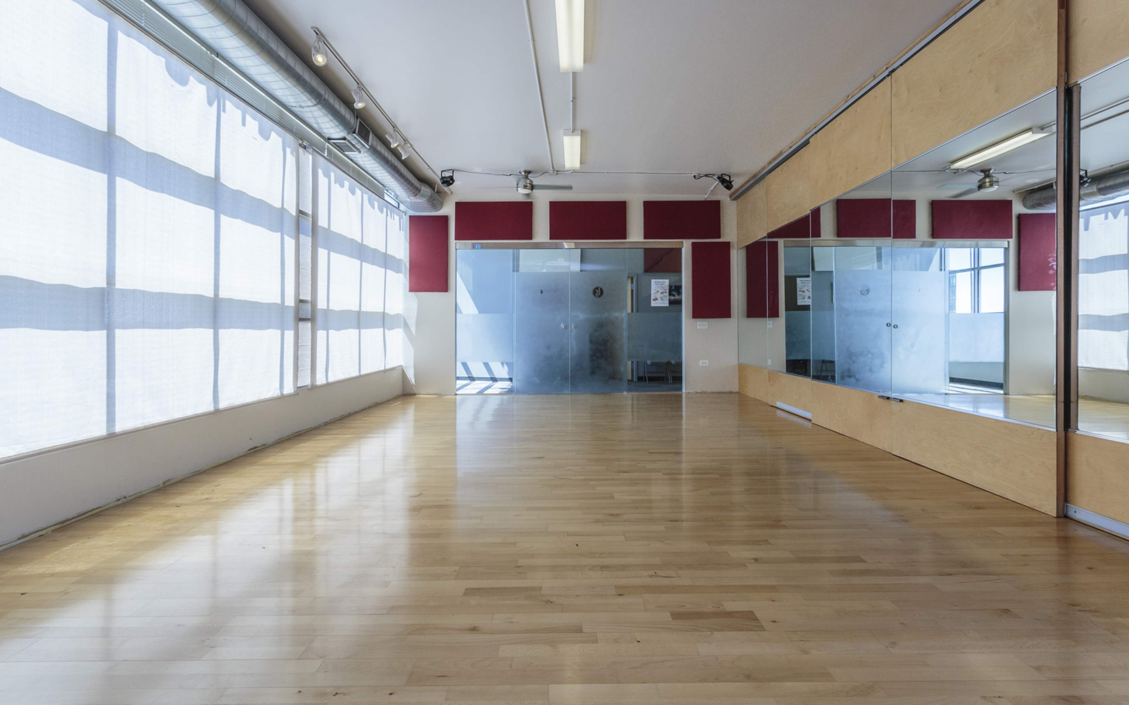 A spacious, empty room with wooden floors, large windows covered by sheer curtains, and a wall of mirrors.
