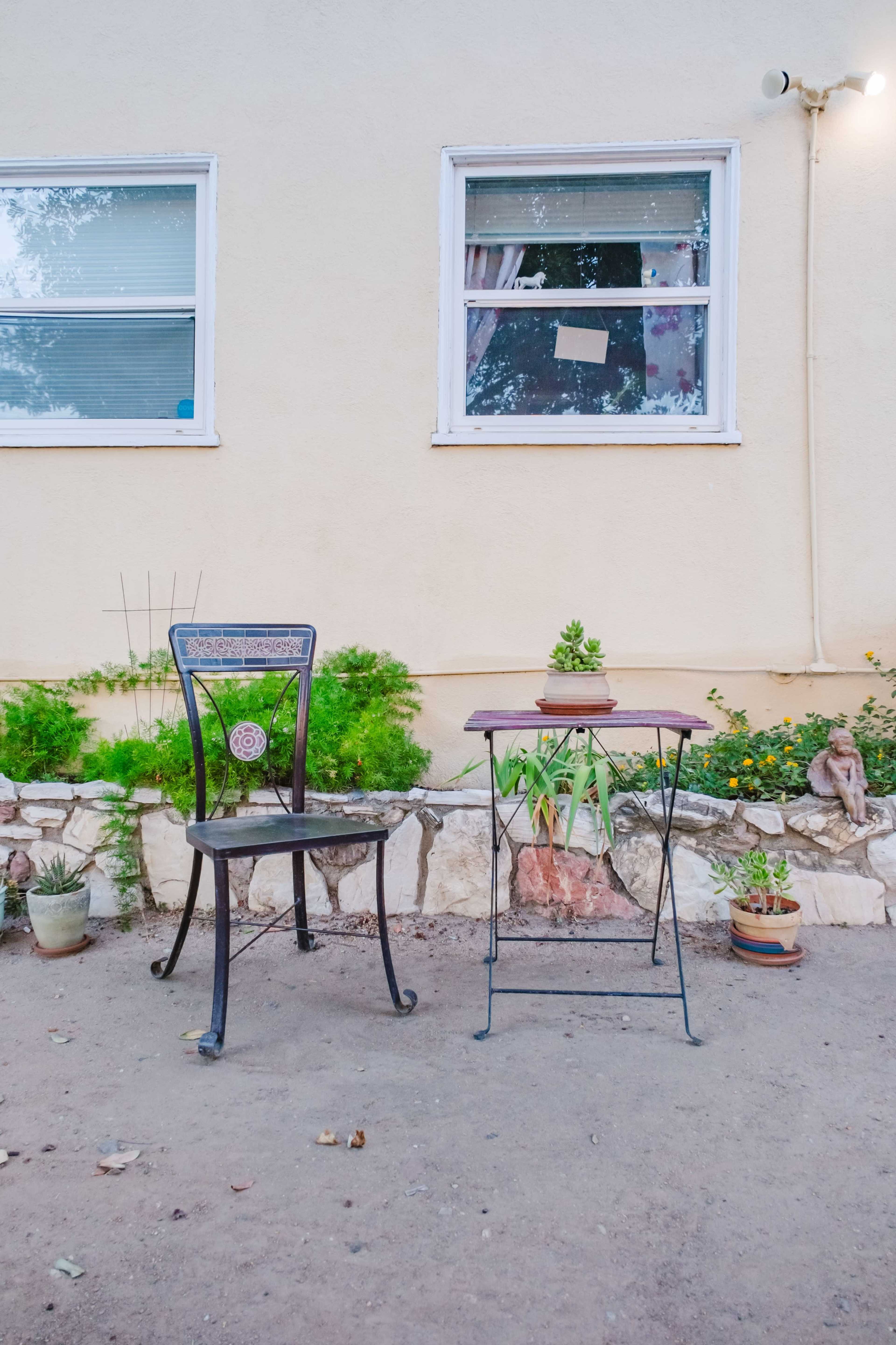 A metal chair and a small table with succulents are placed on a dirt surface next to a light-colored wall with a window.