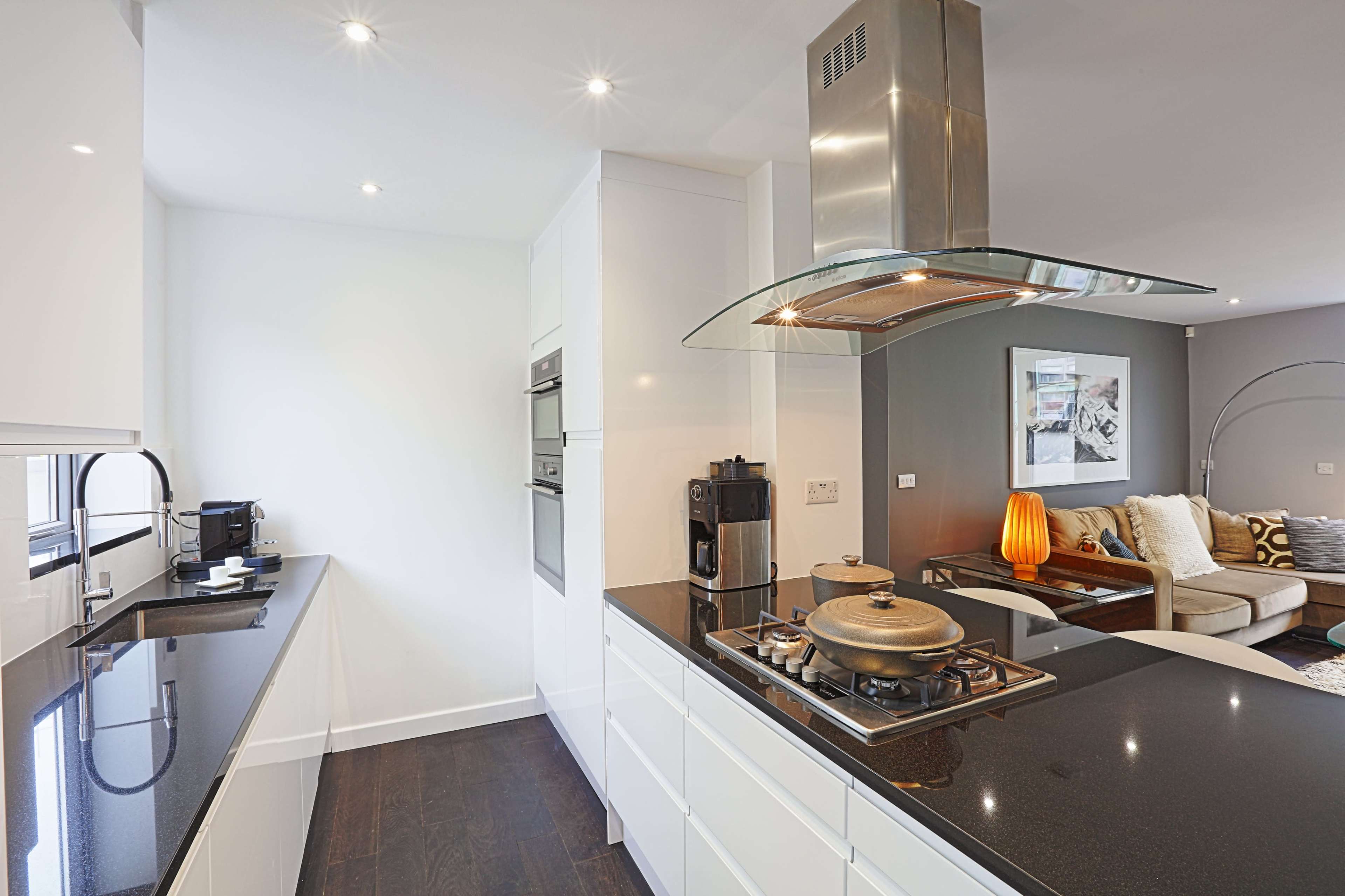 A modern kitchen features sleek white cabinetry, a black countertop, and stainless steel appliances, with a living area visible in the background.