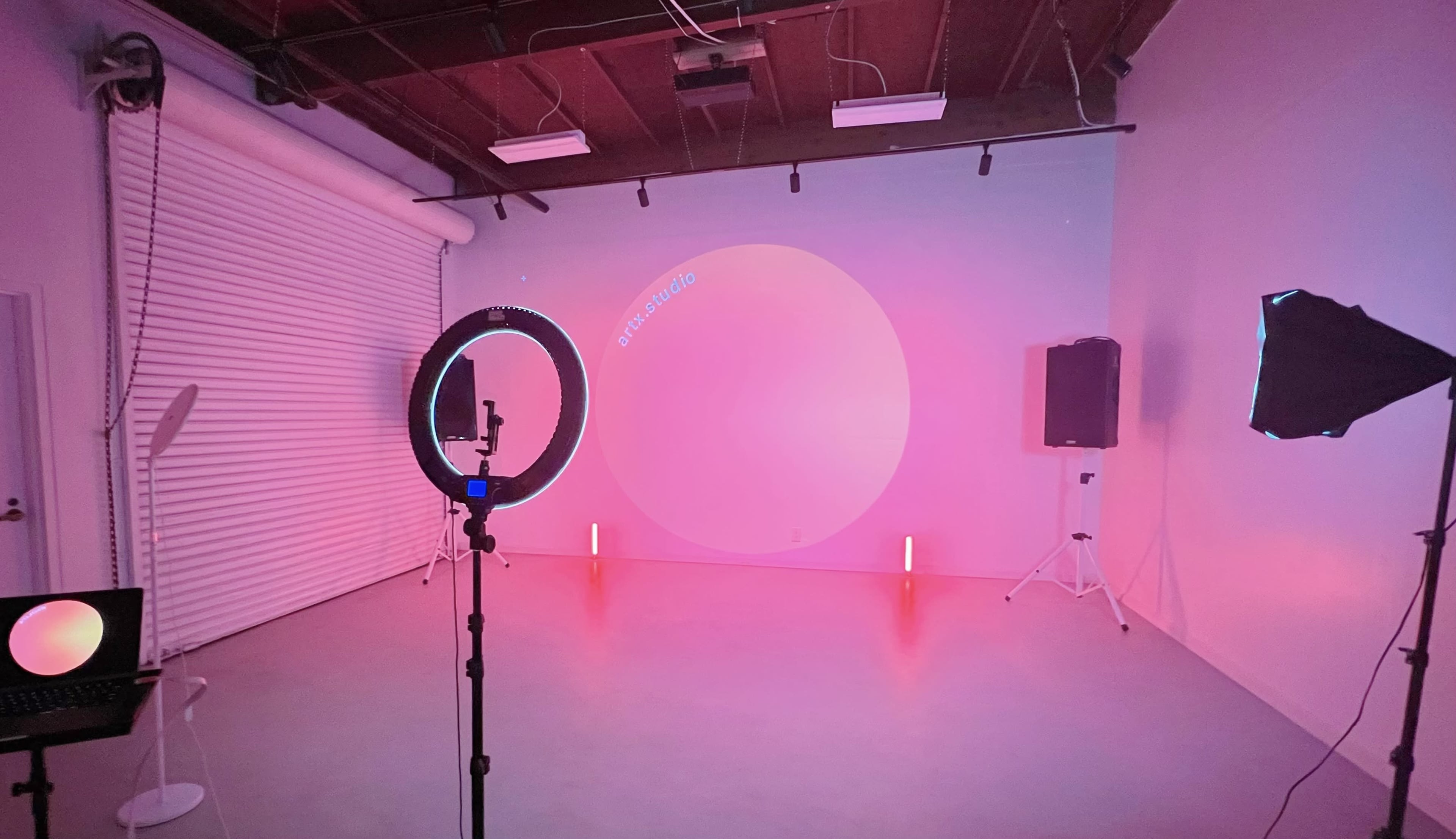 The image shows a brightly lit studio space with a white backdrop, circular light, a camera setup on a tripod, and speakers arranged for a photoshoot or video production.