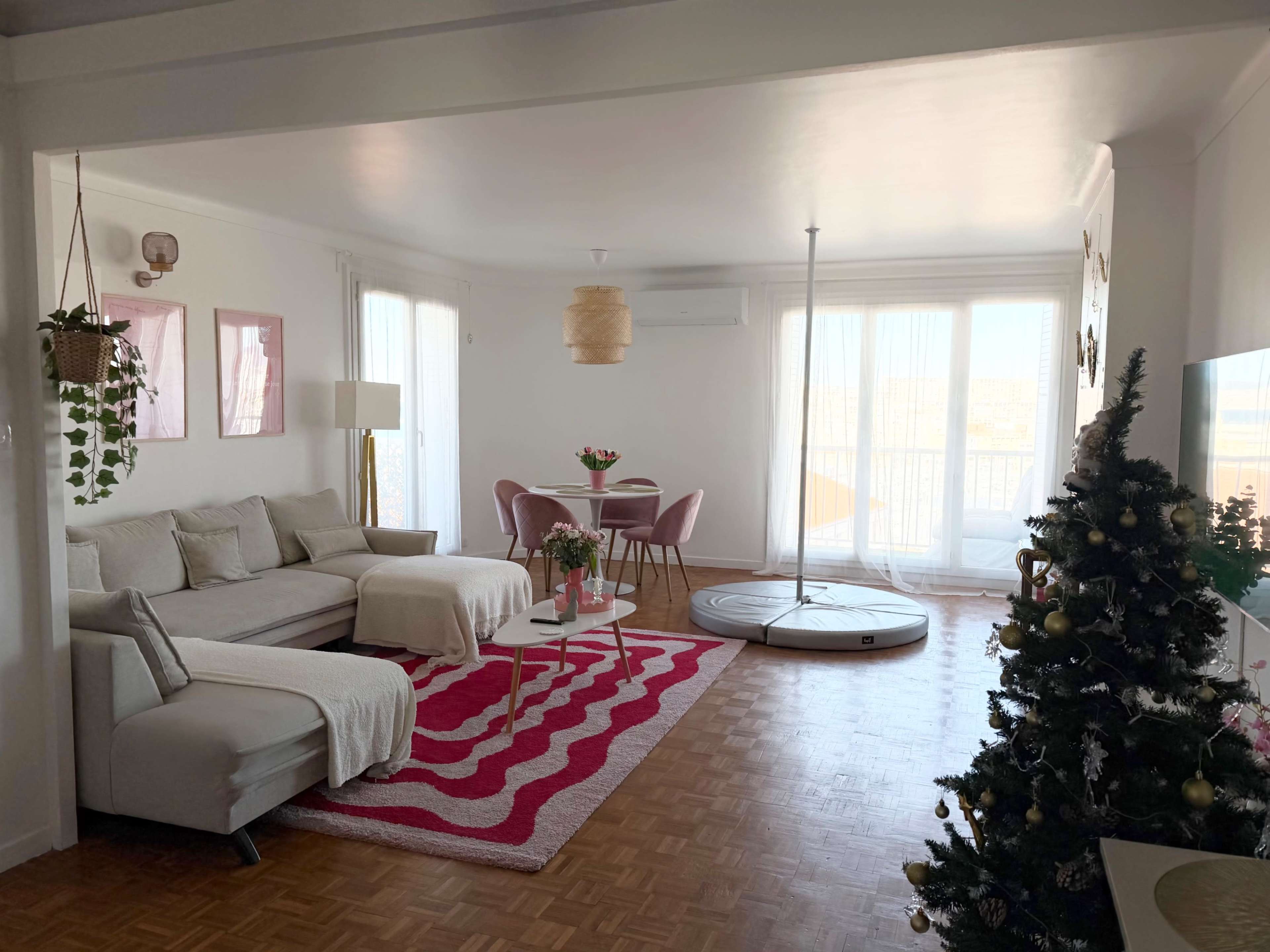 A bright living room features a sectional sofa, a round dining table with pink chairs, and a small Christmas tree decorated with ornaments.