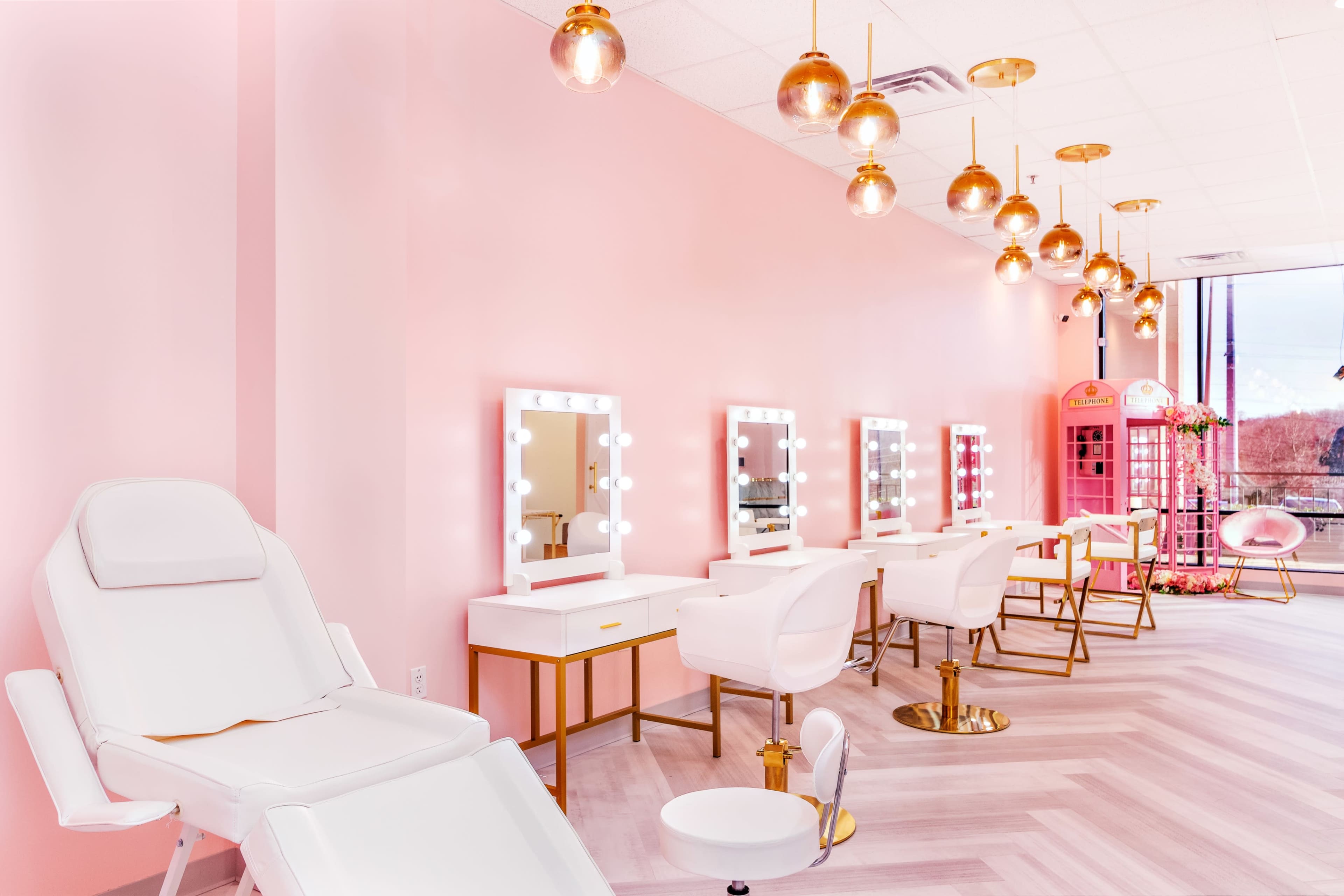 The image depicts a salon interior with pink walls, gold lighting fixtures, and several makeup stations featuring illuminated mirrors.