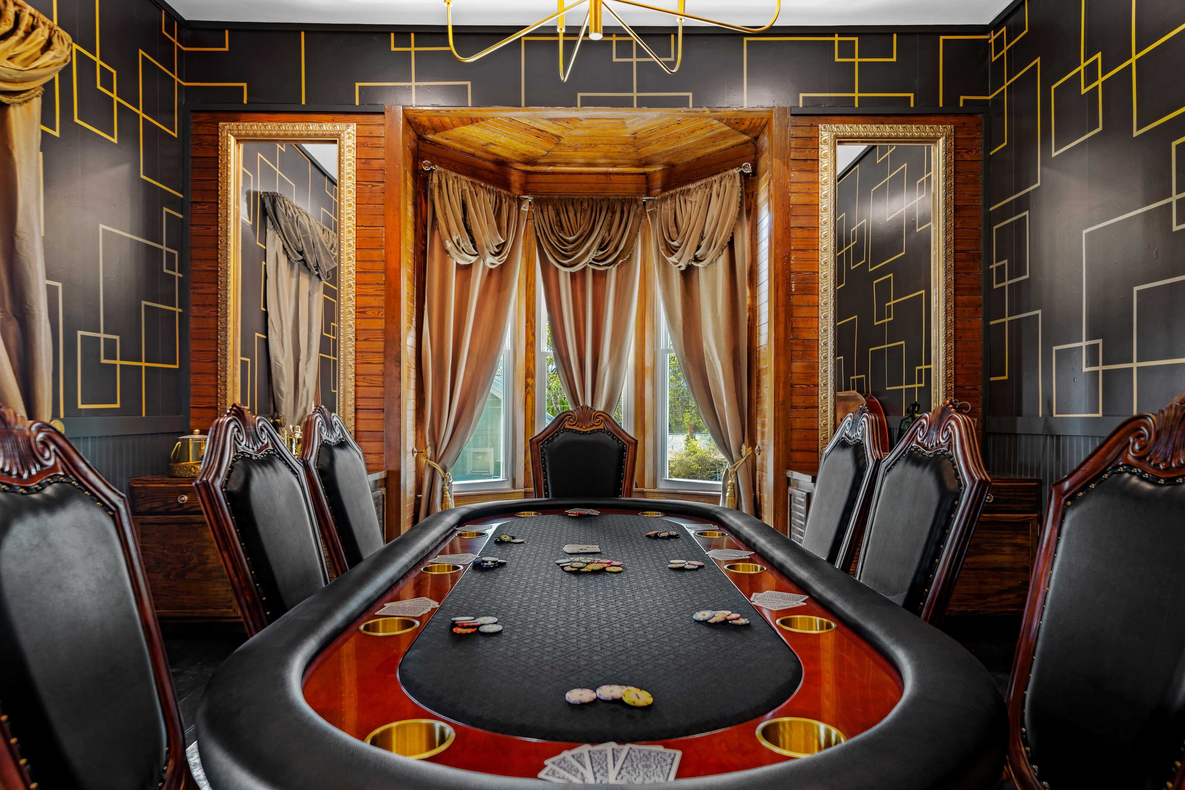A poker room with a large oval table, surrounded by black leather chairs, set against a backdrop of patterned wallpaper and elegant drapes.