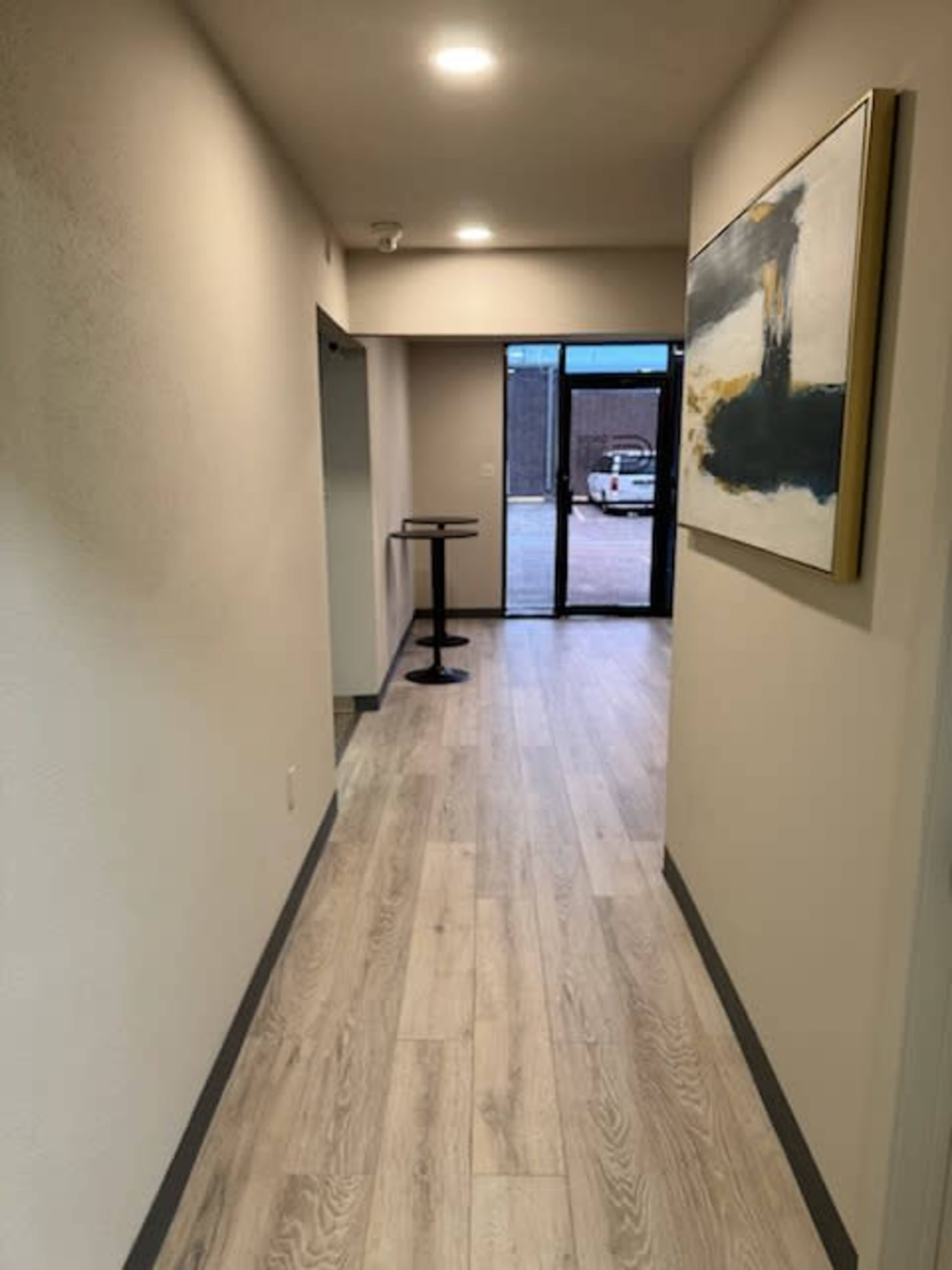 The image shows a well-lit hallway with light-colored flooring and a large painting on one side, leading to a glass door at the end.