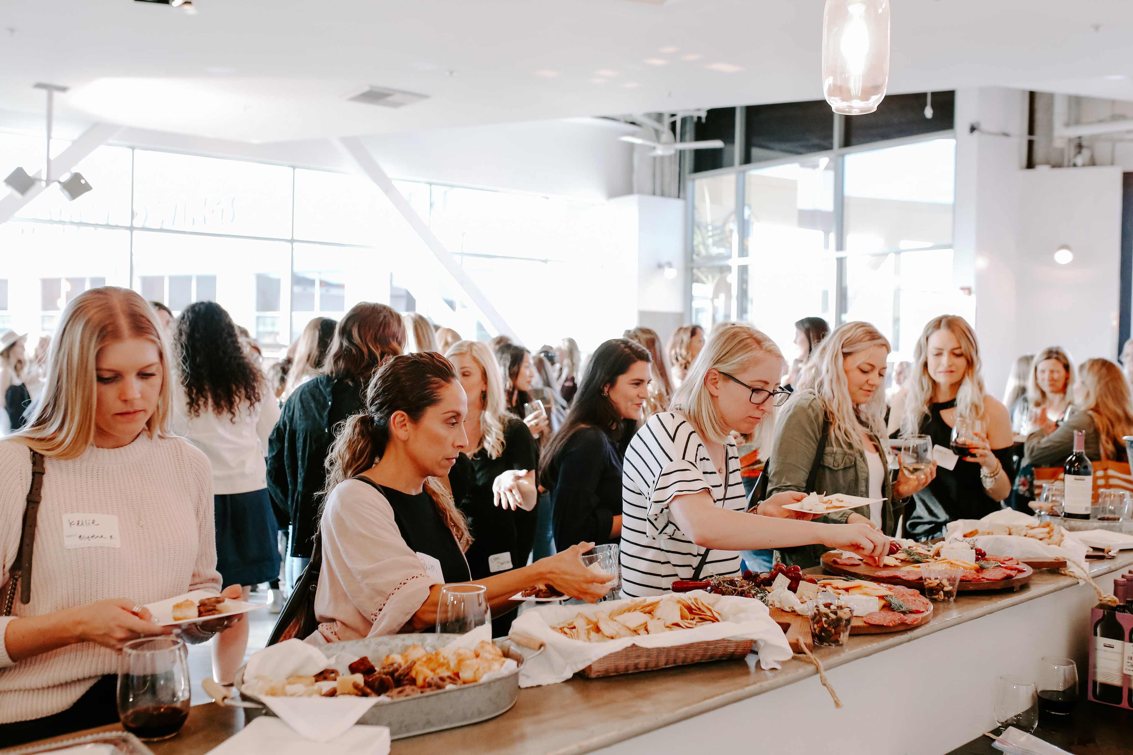 A crowded indoor event features a group of women gathering around a buffet table filled with various food items.