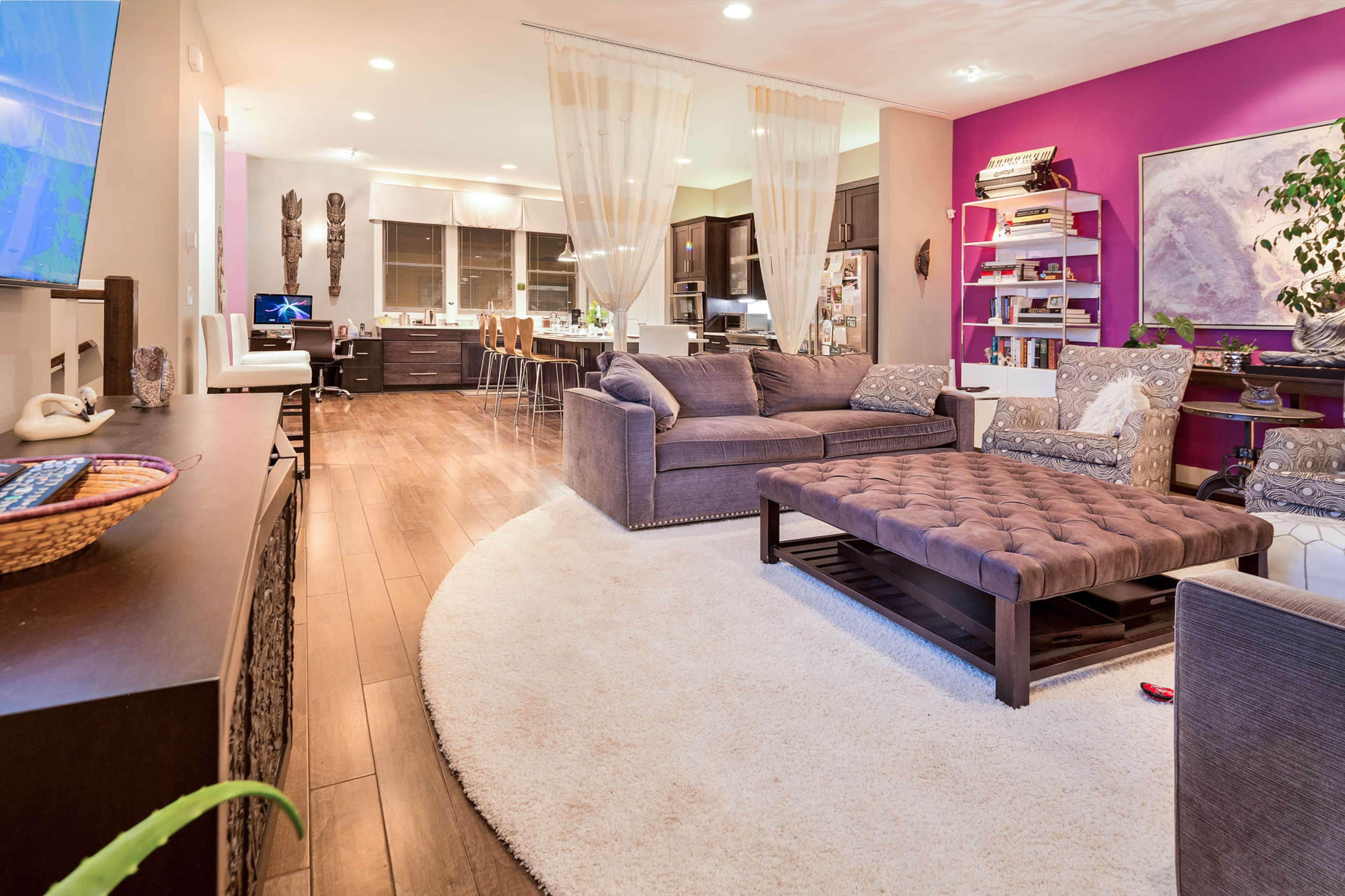 The image shows a modern living room with a sectional couch, a coffee table, and a purple accent wall, alongside a kitchen area and decorative elements.