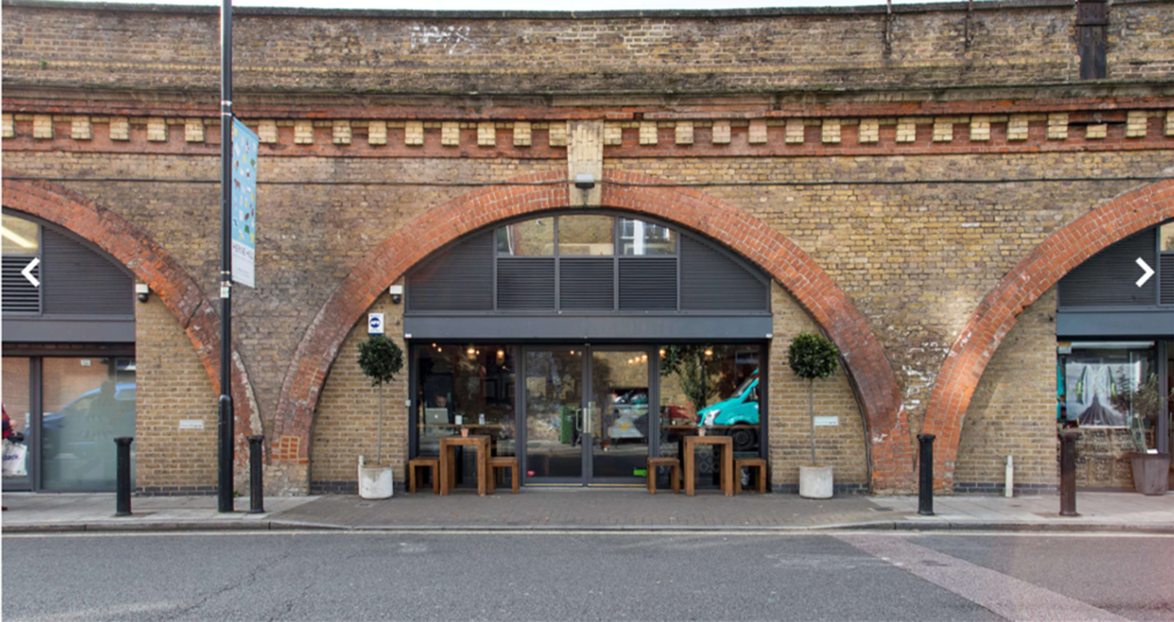 The entrance of a brick building with large arched windows, flanked by two small tables outside, under a railway arch.
