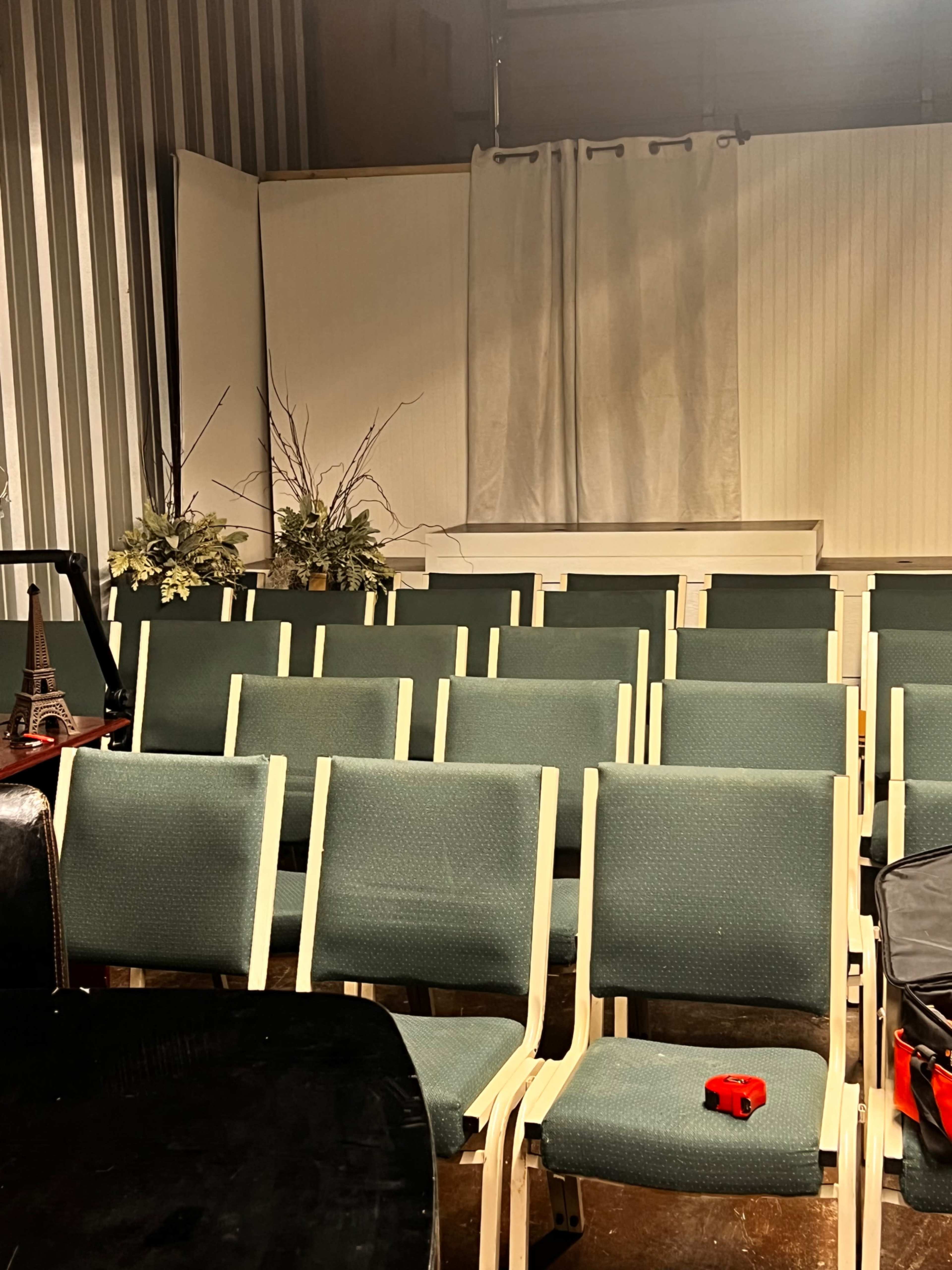 The image shows a row of empty green chairs arranged in a room featuring a backdrop with a curtain and a decorative arrangement of foliage.