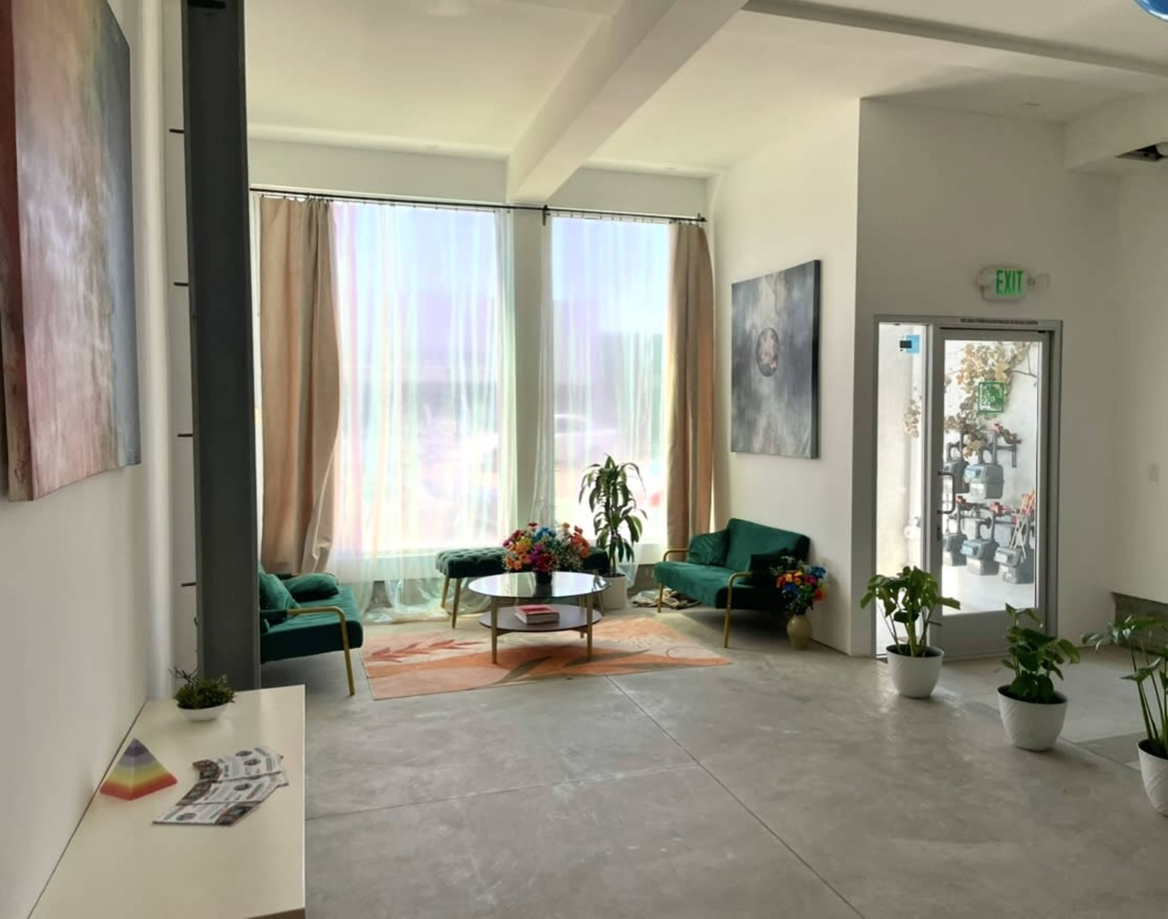 The image shows a modern interior space with large windows, green seating, a coffee table, and potted plants.