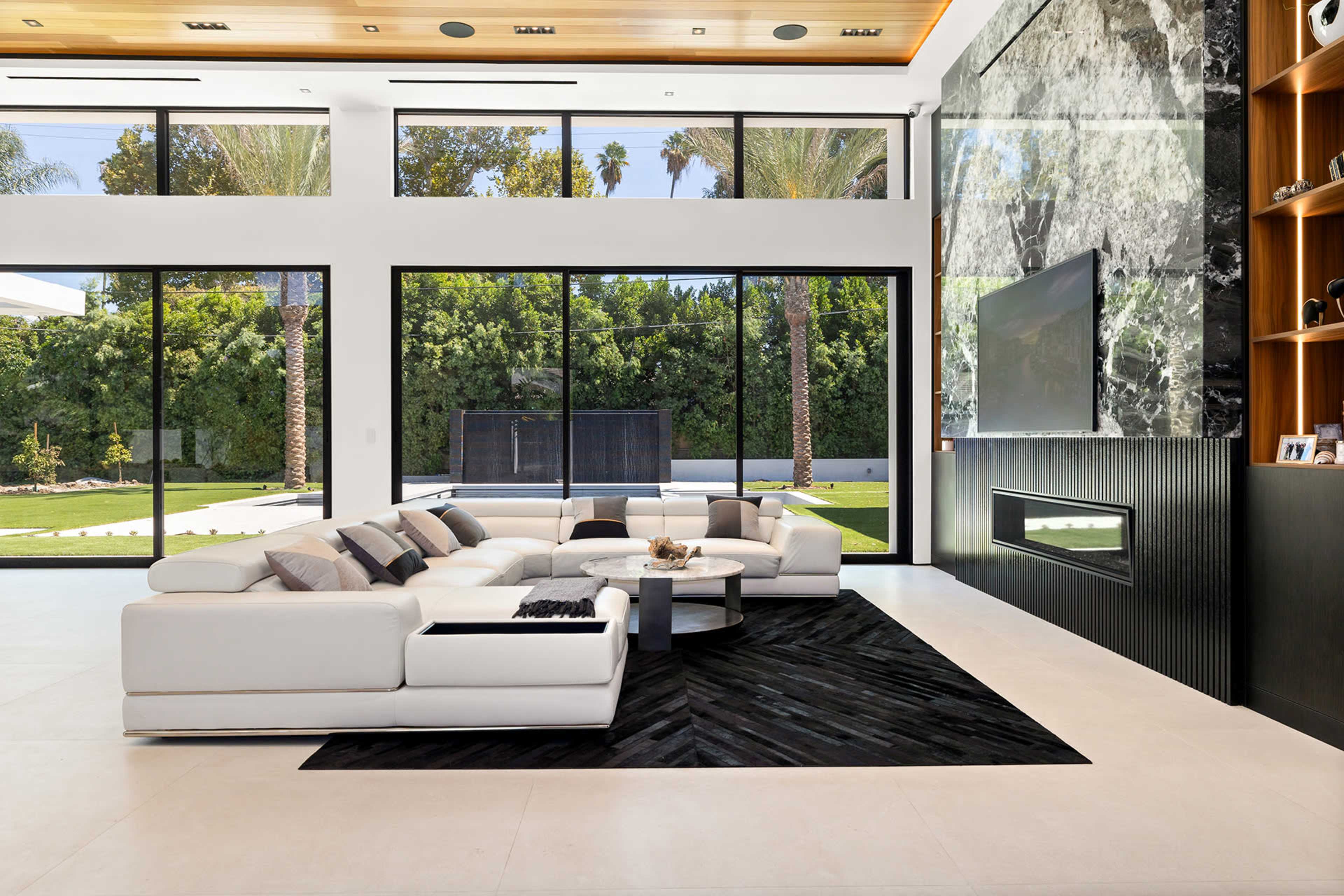 A modern living room features a white sectional sofa, a circular coffee table, and large glass windows overlooking a landscaped area with palm trees.