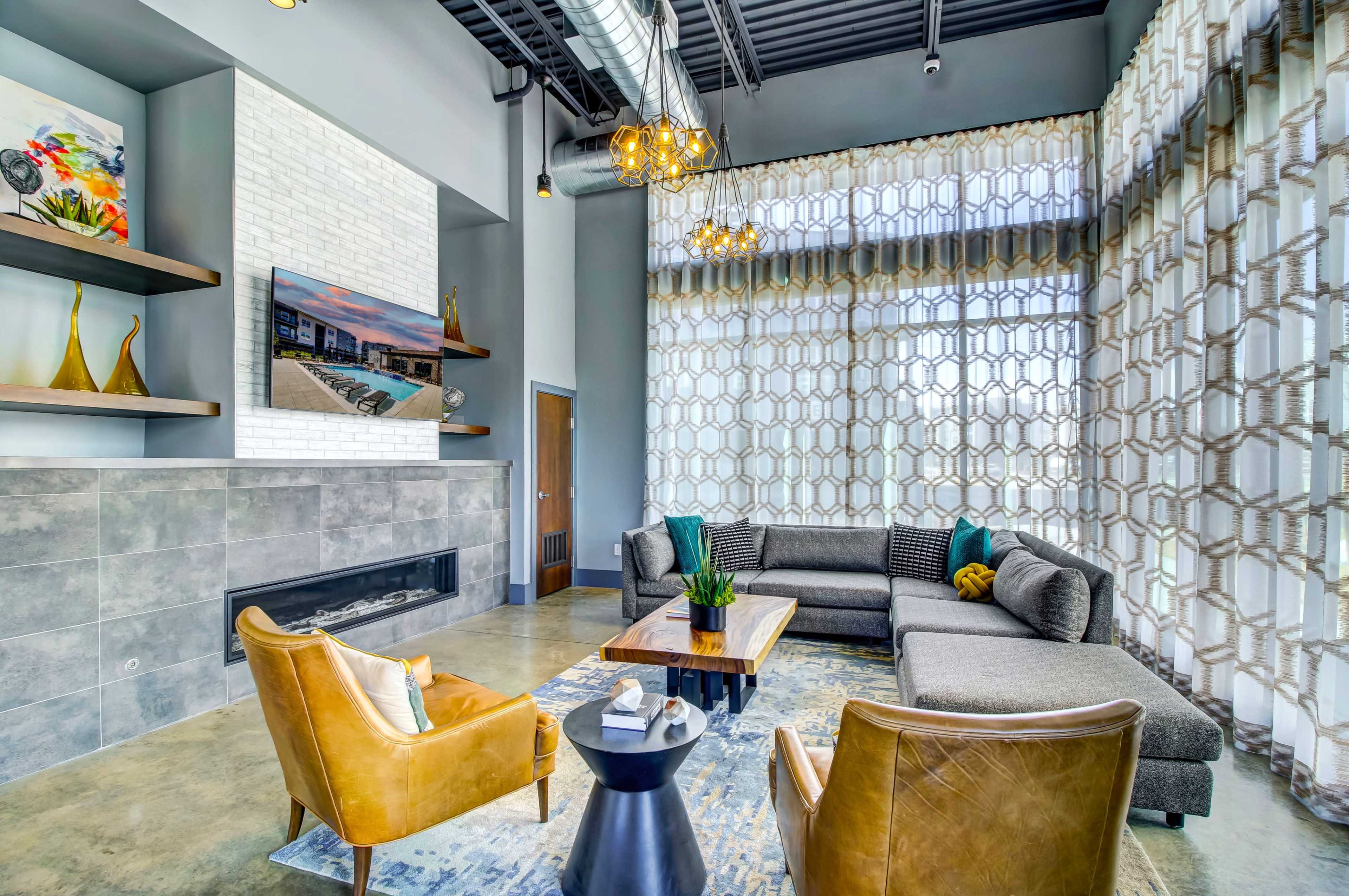 A modern living area featuring a sectional sofa, two armchairs, a coffee table, and large windows with patterned drapes, illuminated by overhead lighting.