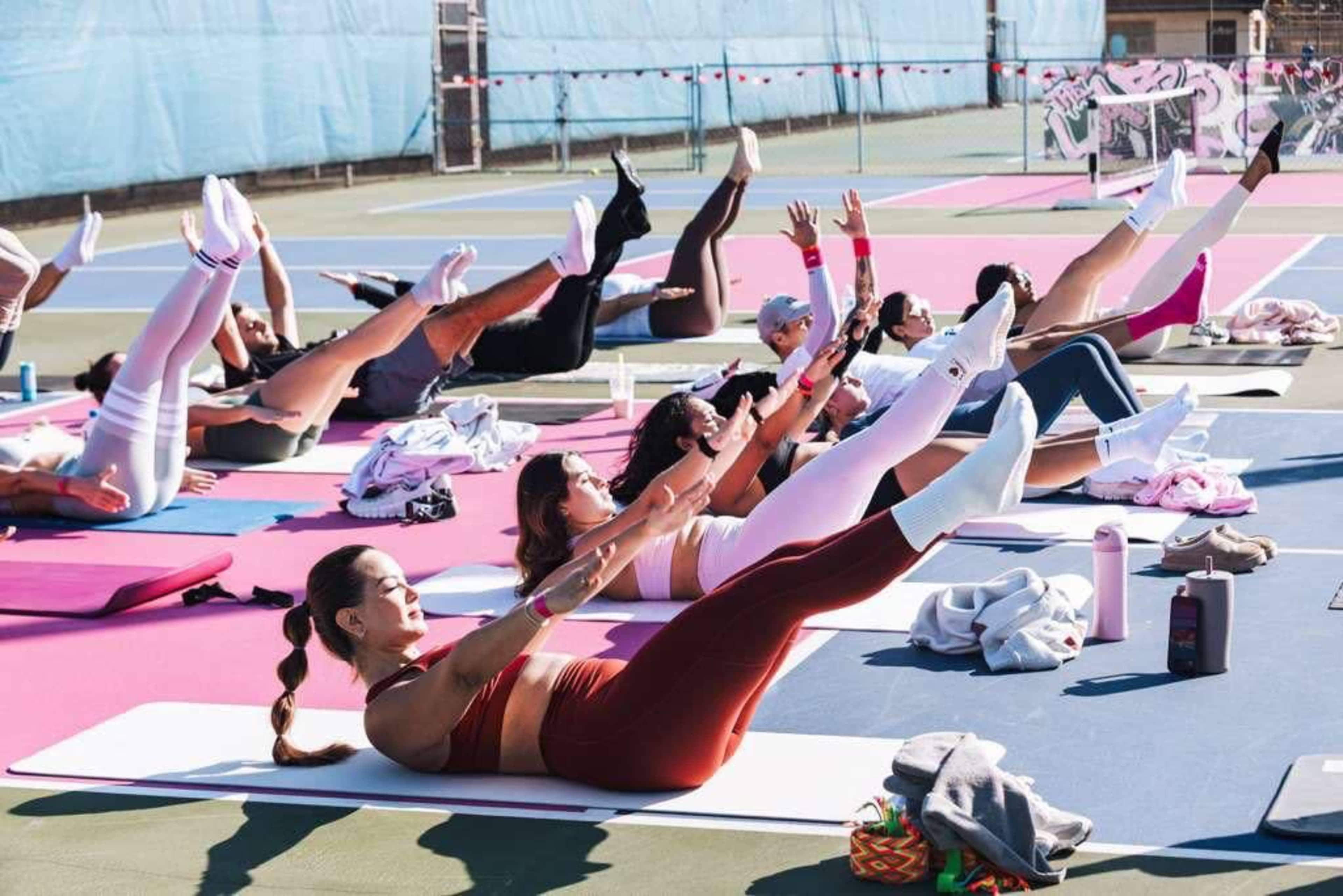 A group of individuals participates in a fitness class on colorful mats, performing leg lifts on a tennis court.