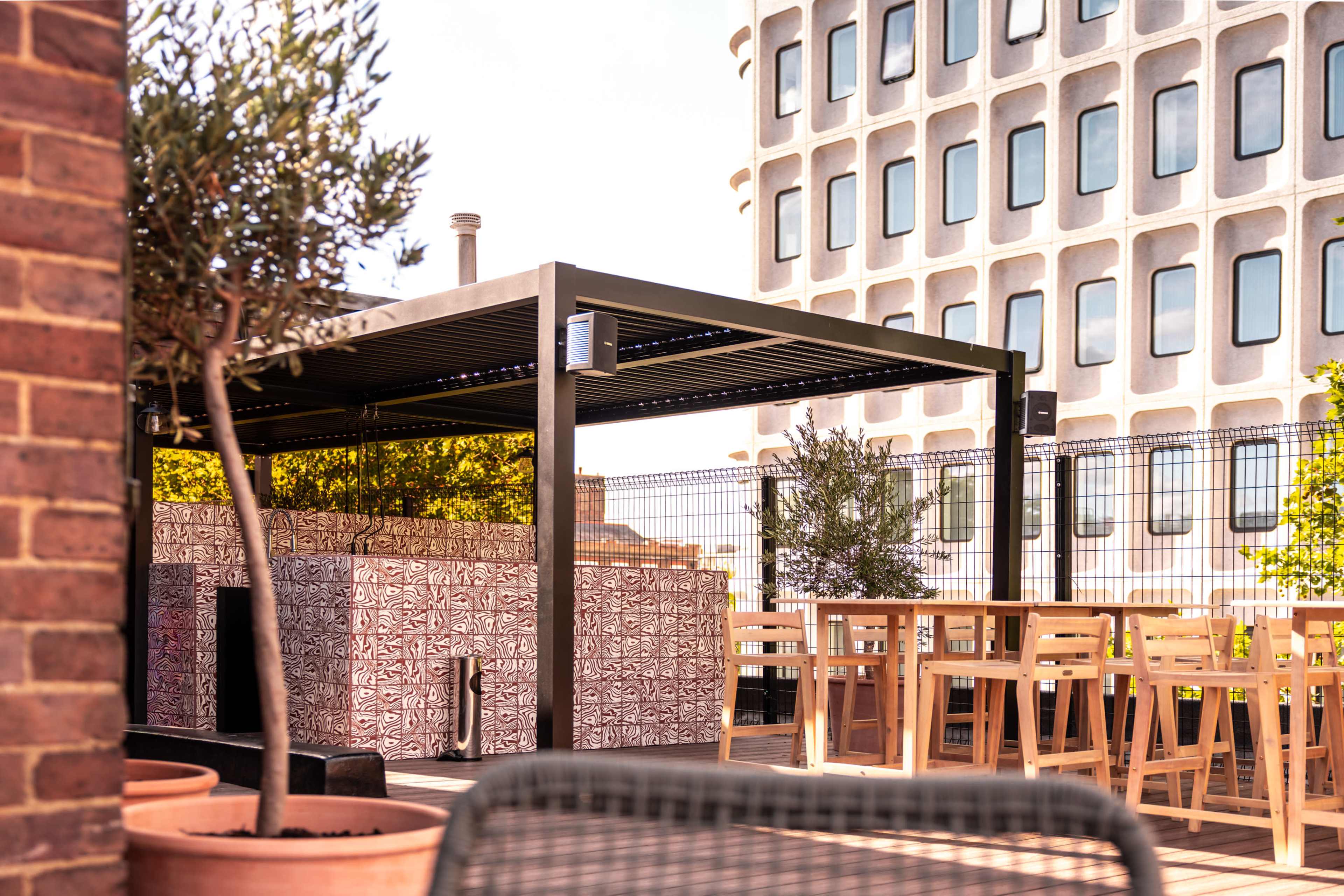 A rooftop terrace features a shaded seating area with a decorative wall, surrounded by greenery and adjacent to a modern building.