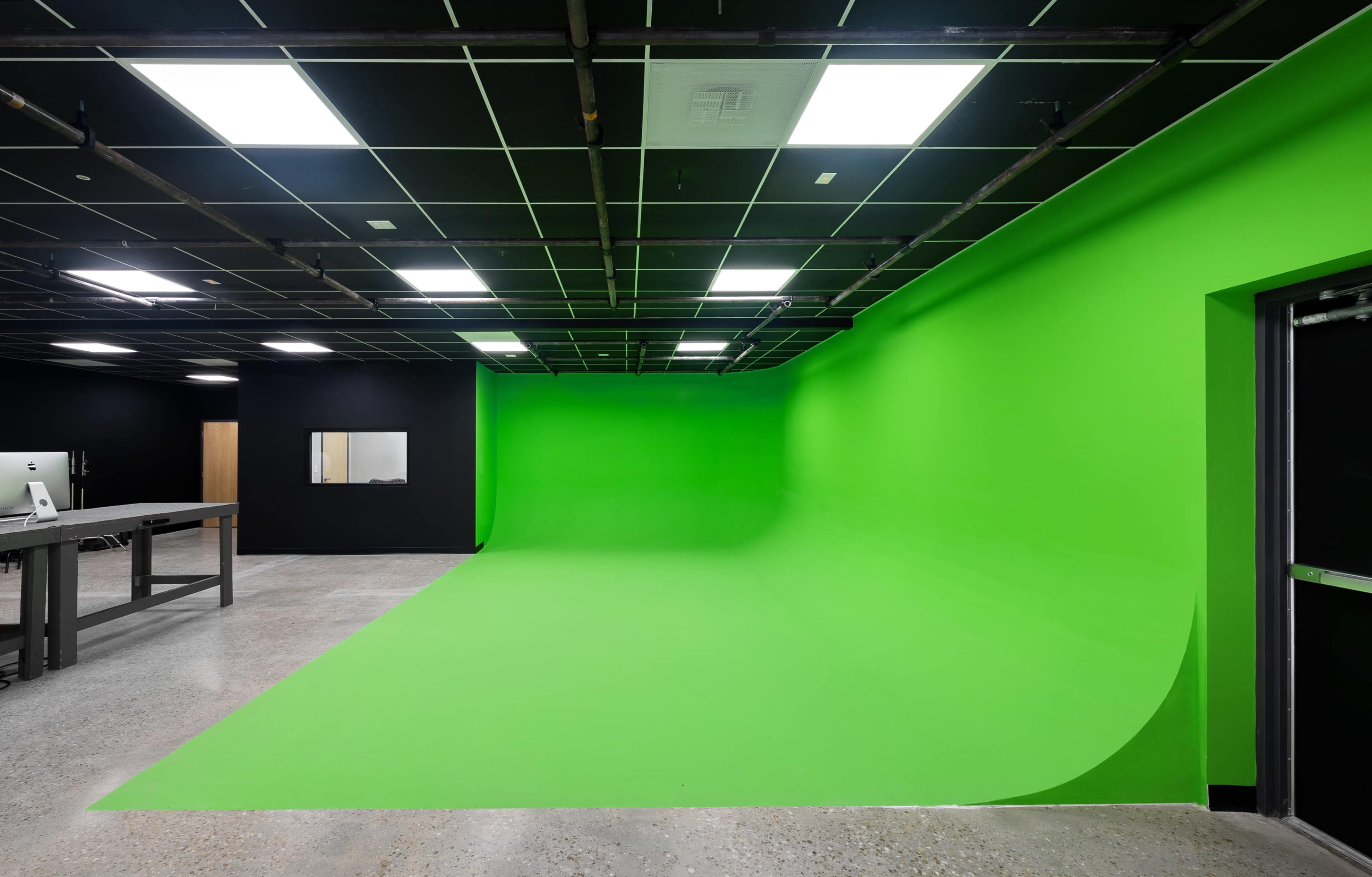A spacious studio with a large green screen on one wall and black walls, featuring overhead lighting and a workbench on one side.
