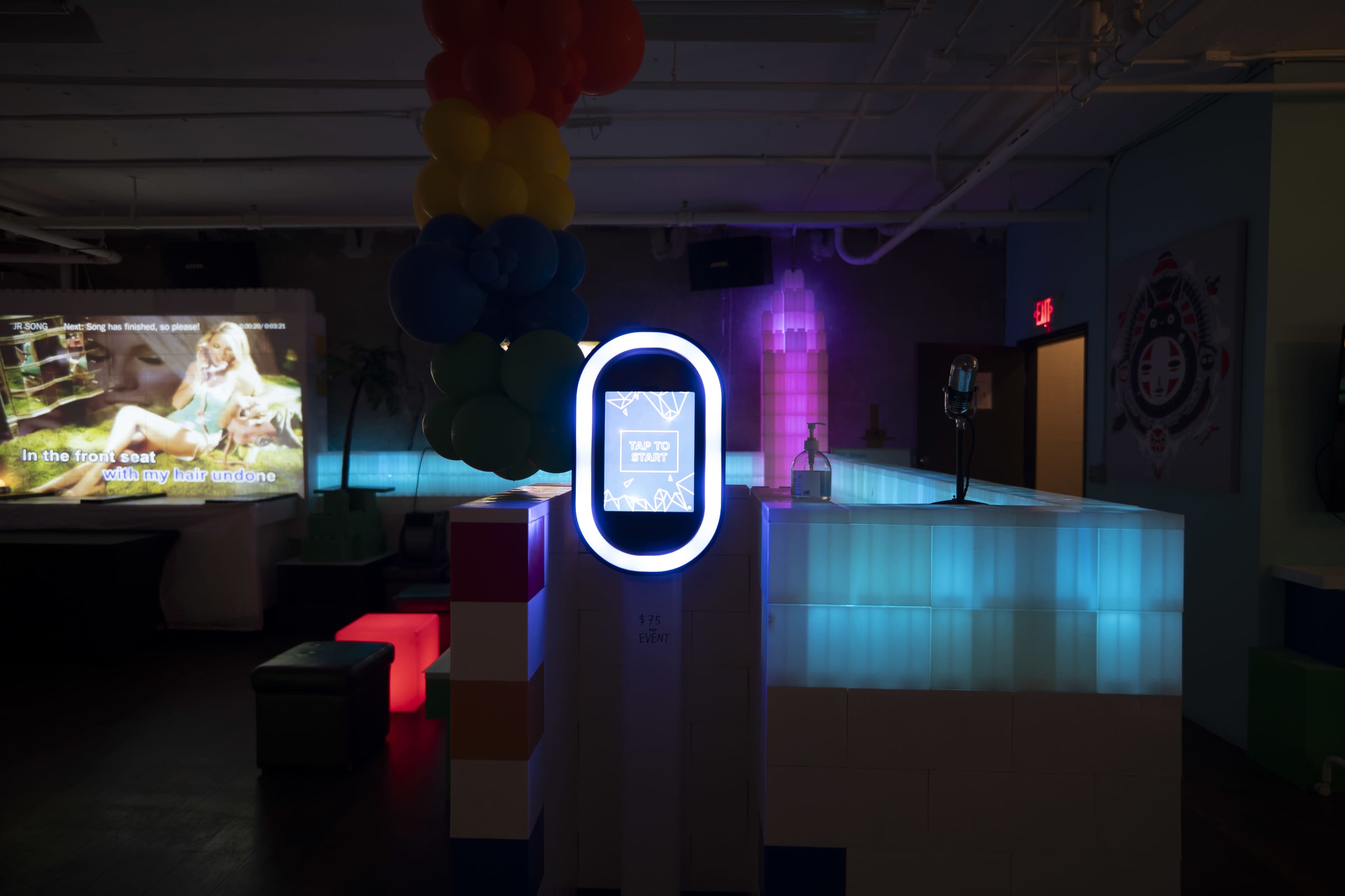 A brightly lit bar area featuring a digital screen, colorful balloons, and a neon sign indicating a tap-to-start activation.