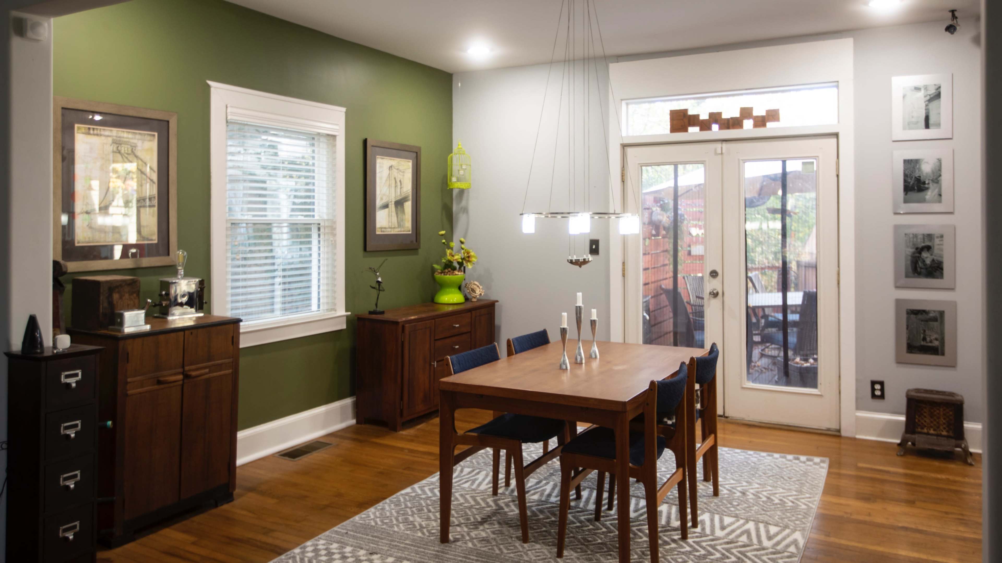 A dining room features a wooden table surrounded by chairs, a sideboard, and artwork on the walls, with a door leading to an outdoor area.
