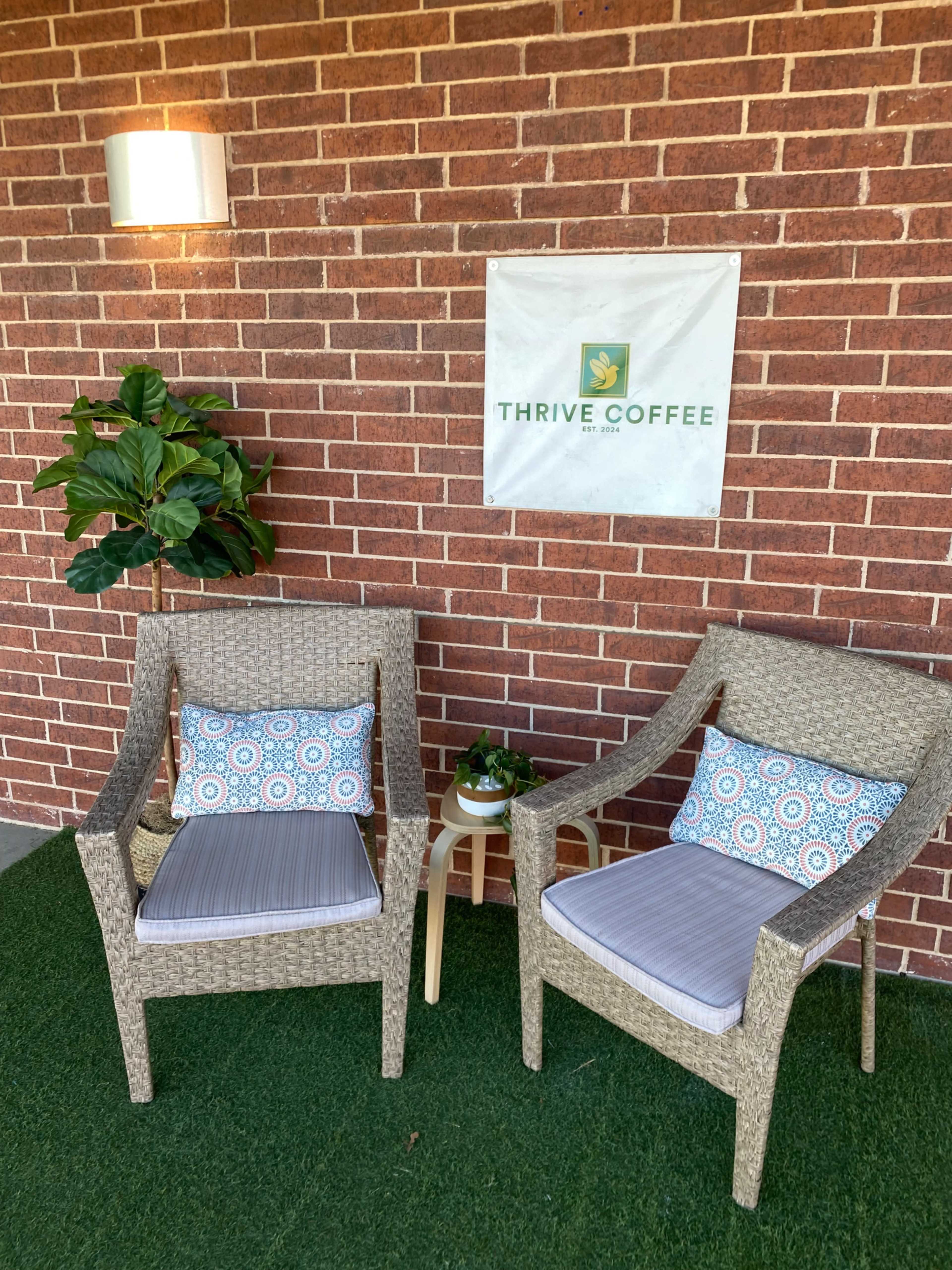 The image shows two wicker chairs with cushions, a small table, a potted plant, and a sign for "Thrive Coffee" against a brick wall.