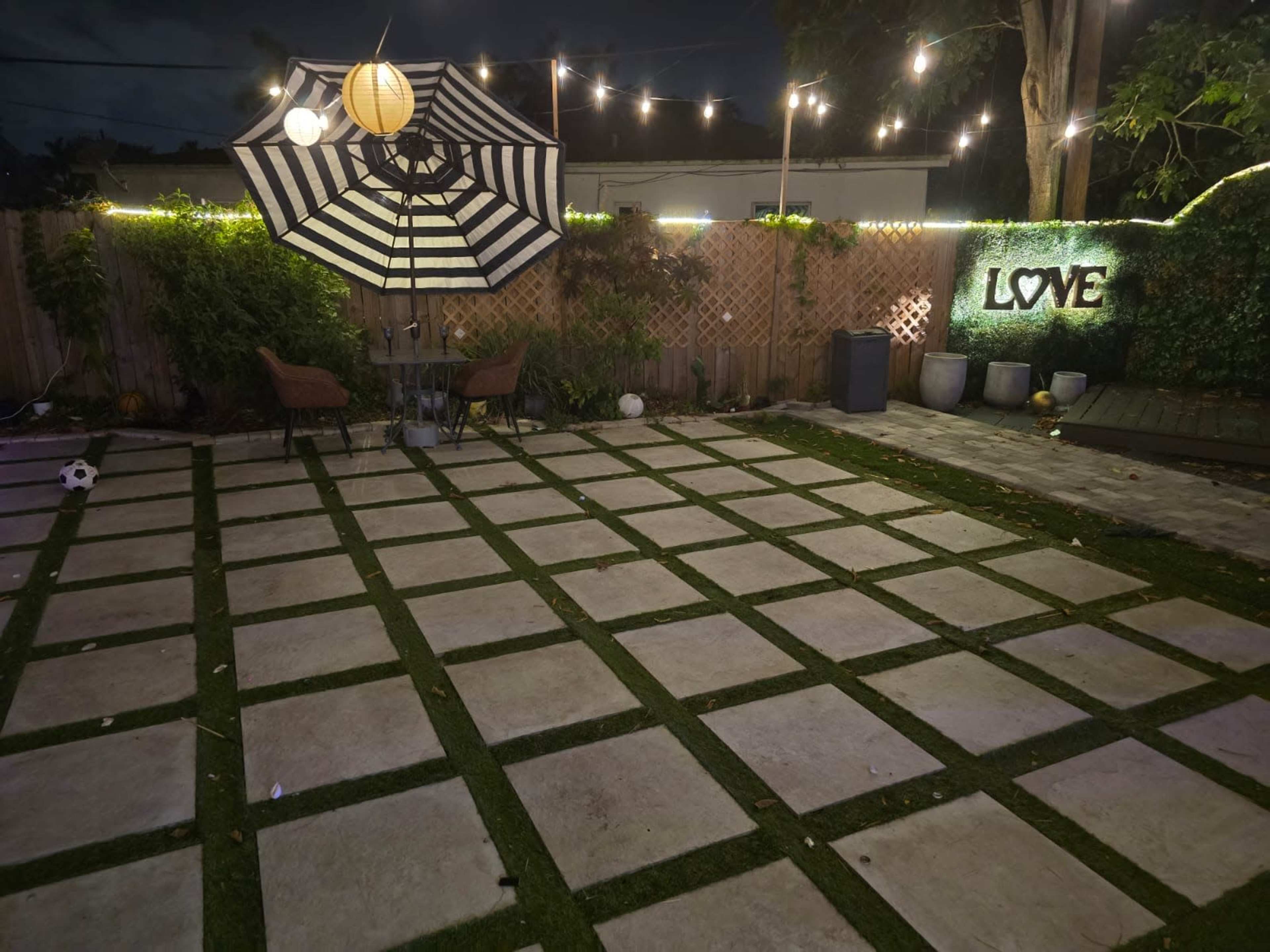 The image shows a backyard patio with geometric stone slabs, grass patches, an umbrella, and decorative lights overhead.