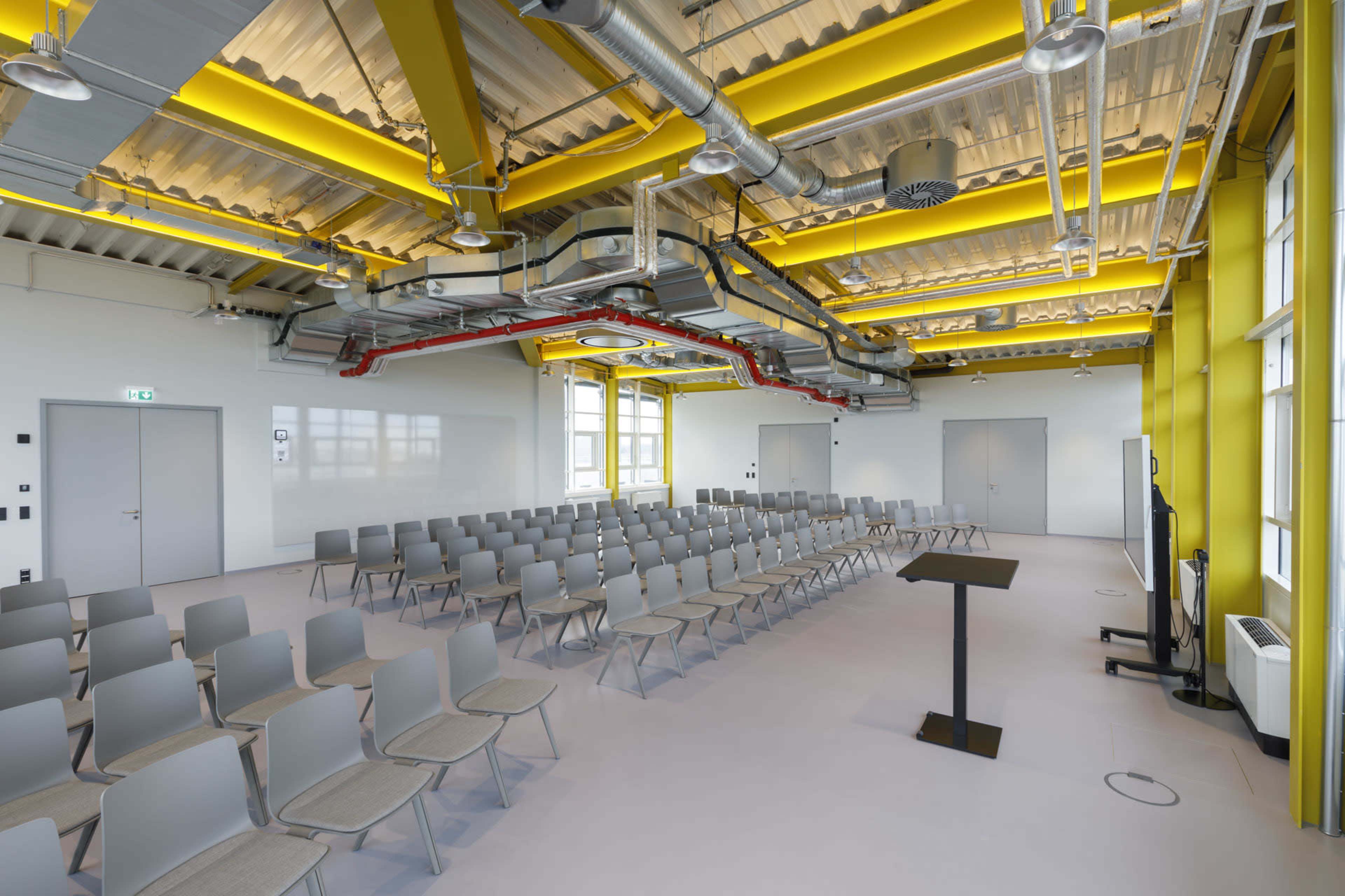 The image shows a modern lecture hall with rows of gray chairs facing a podium, illuminated by yellow and silver industrial-style ceiling elements.