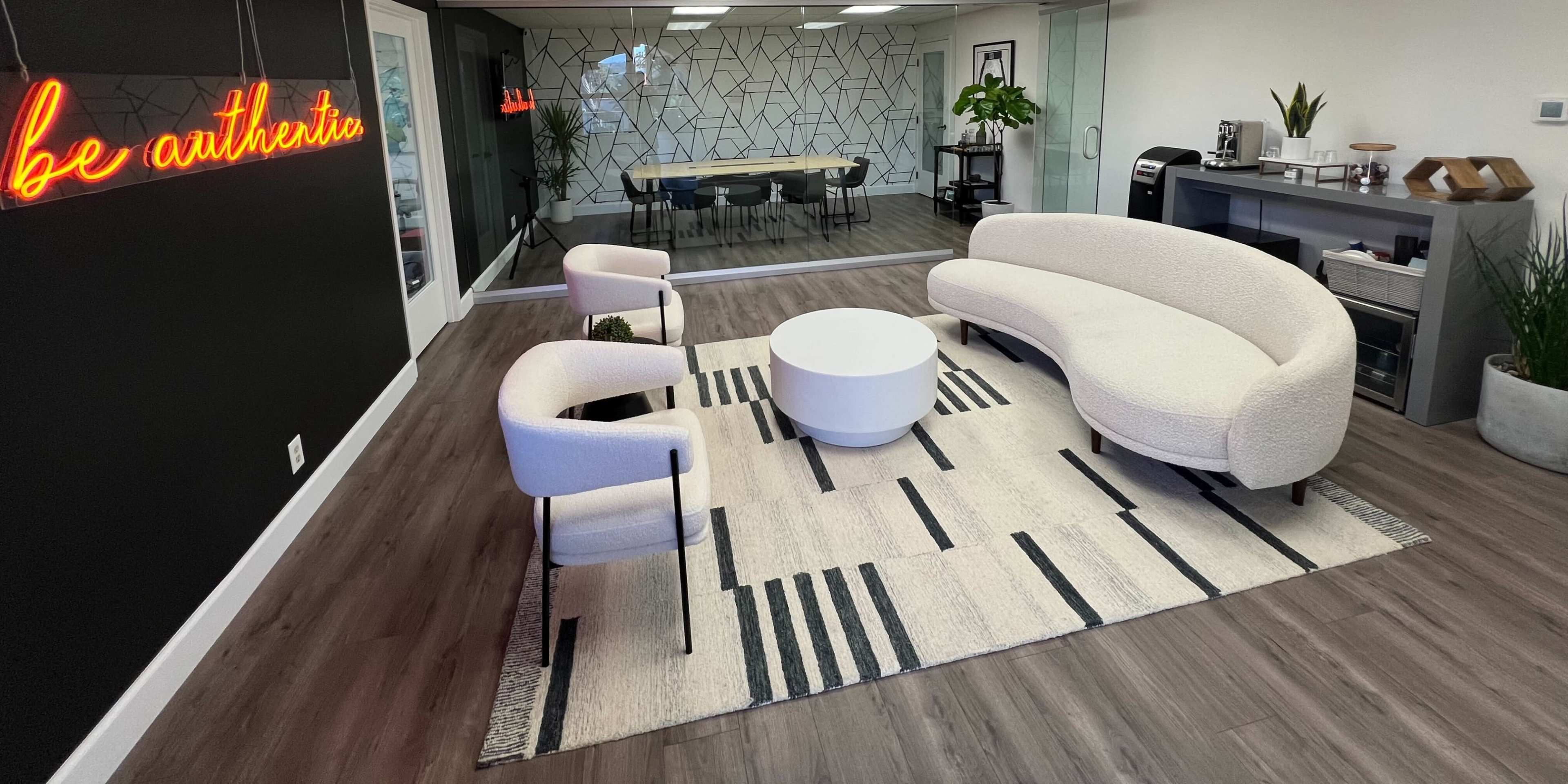 A modern office space featuring a circular white coffee table, a curved white sofa, two white chairs, a patterned area rug, and a glass-walled meeting room in the background.