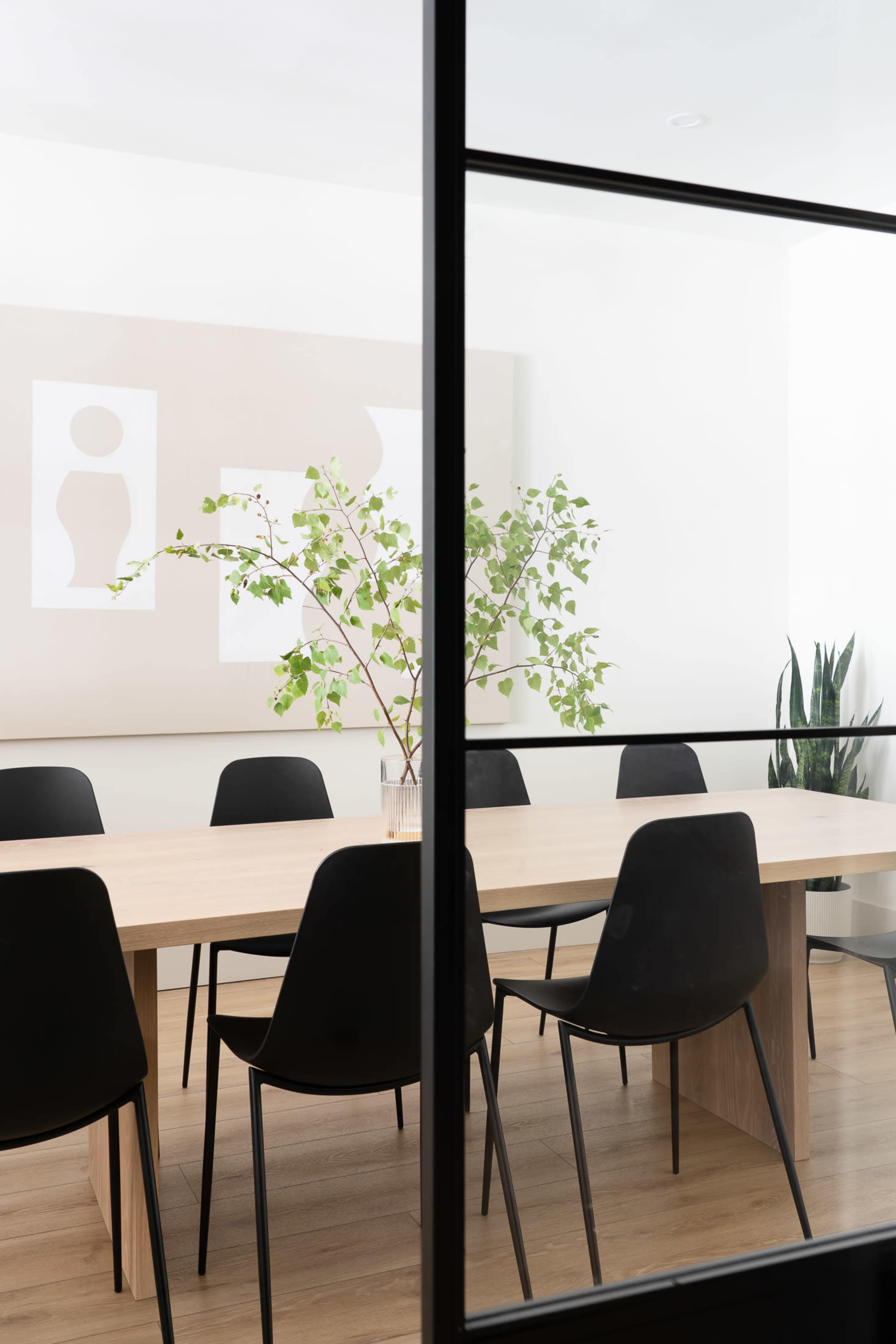 A modern conference room with a large wooden table surrounded by black chairs and a vase of greenery, viewed through a glass door.