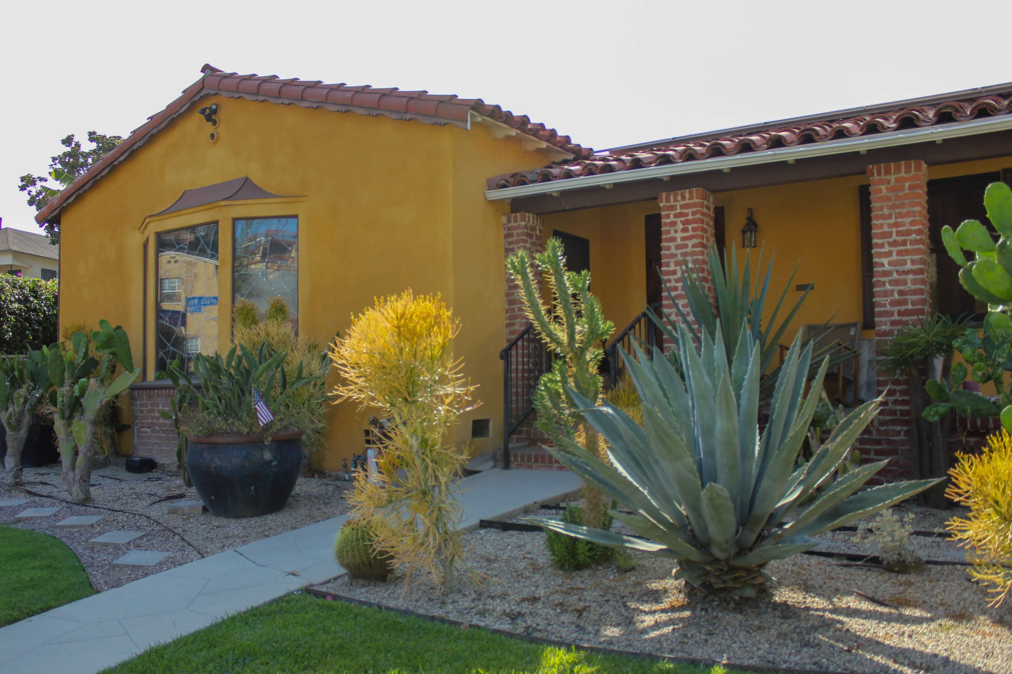 The image shows a colorful house with a yellow exterior and terracotta roof, surrounded by various cacti and desert plants in the front yard.