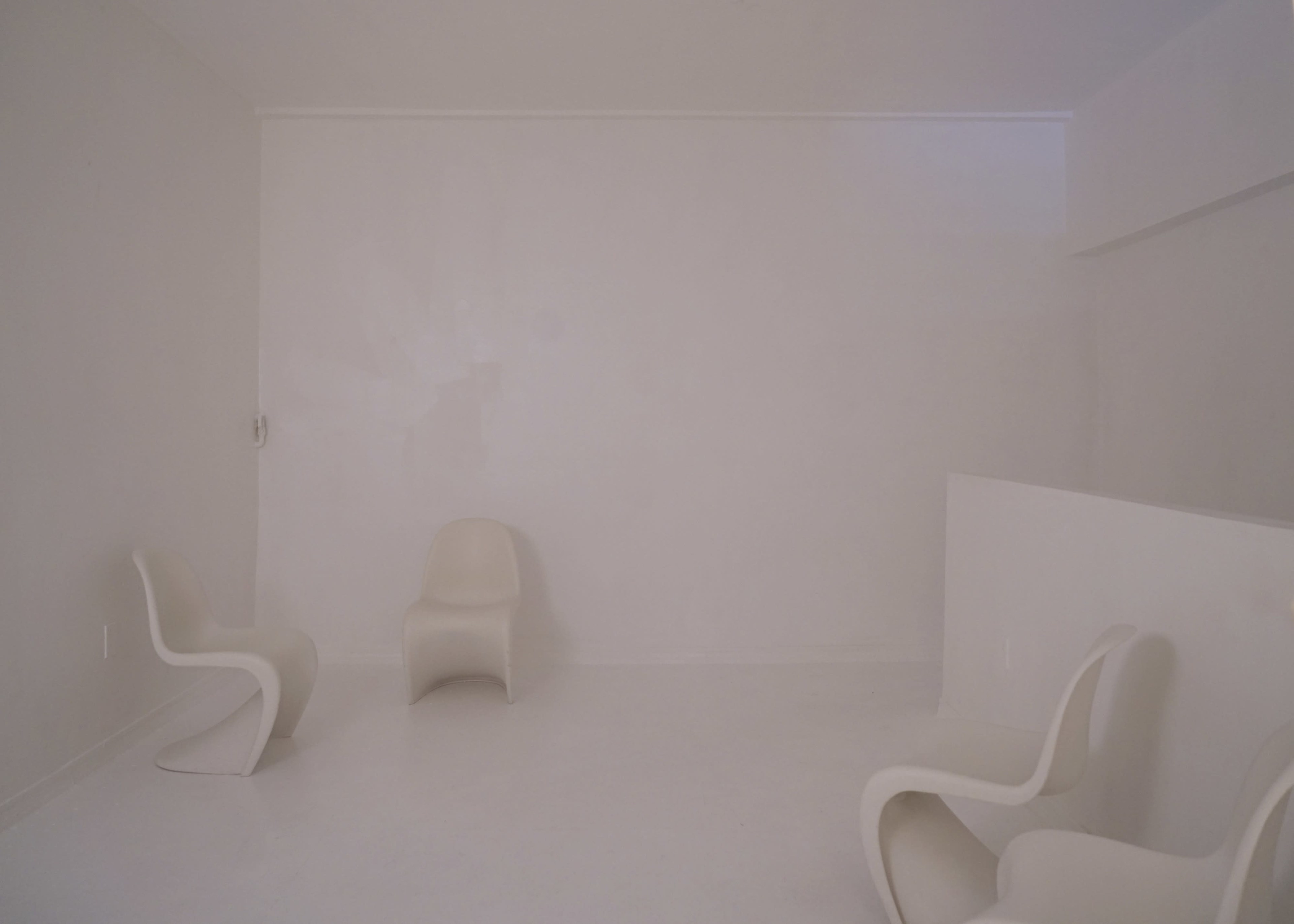 The image shows a minimalist room with white walls and three white chairs arranged on a light-colored floor.