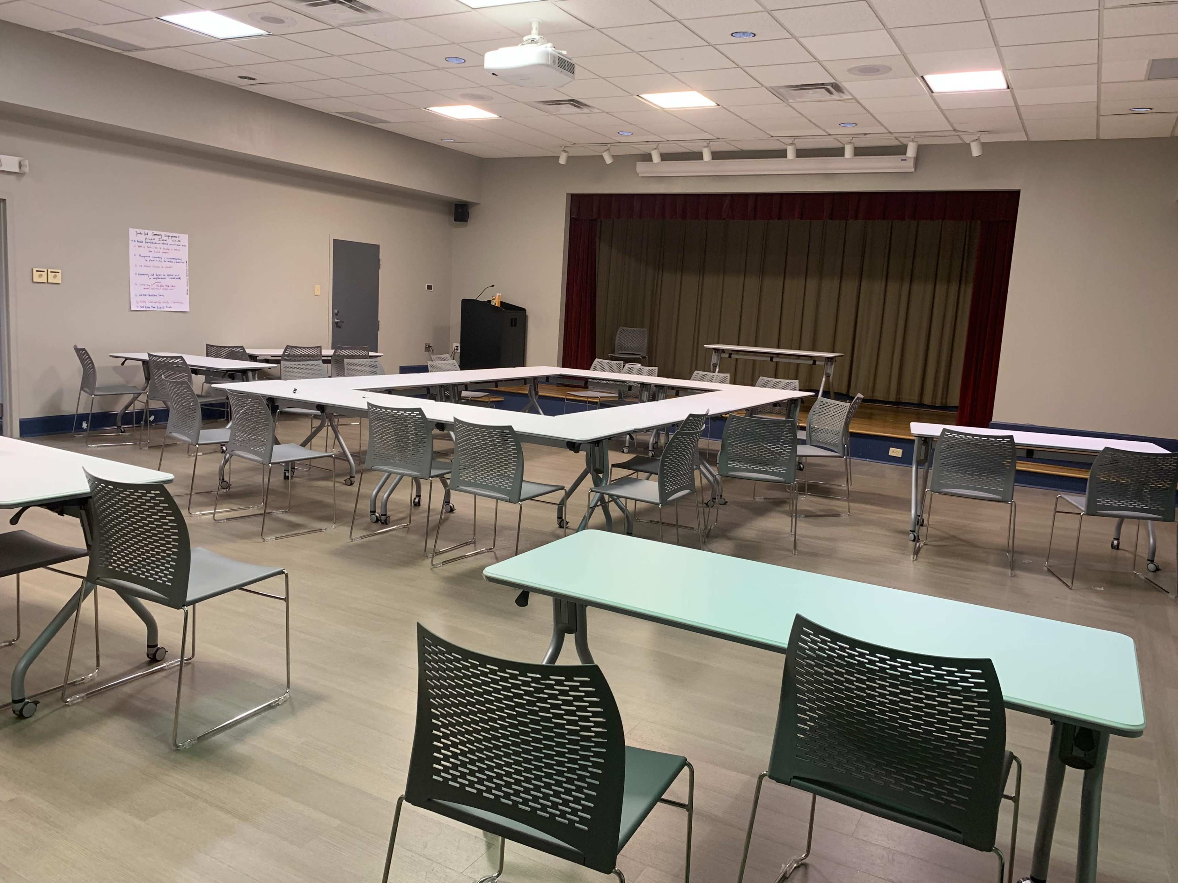 A well-lit meeting room with several tables and chairs arranged in a sparse layout, facing a small stage area.